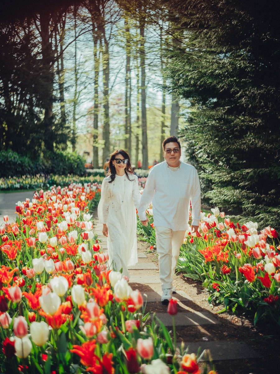 Stel in het wit met zonnebrillen loopt over een pad met rode en witte tulpen, terwijl hoge bomen lange schaduwen werpen