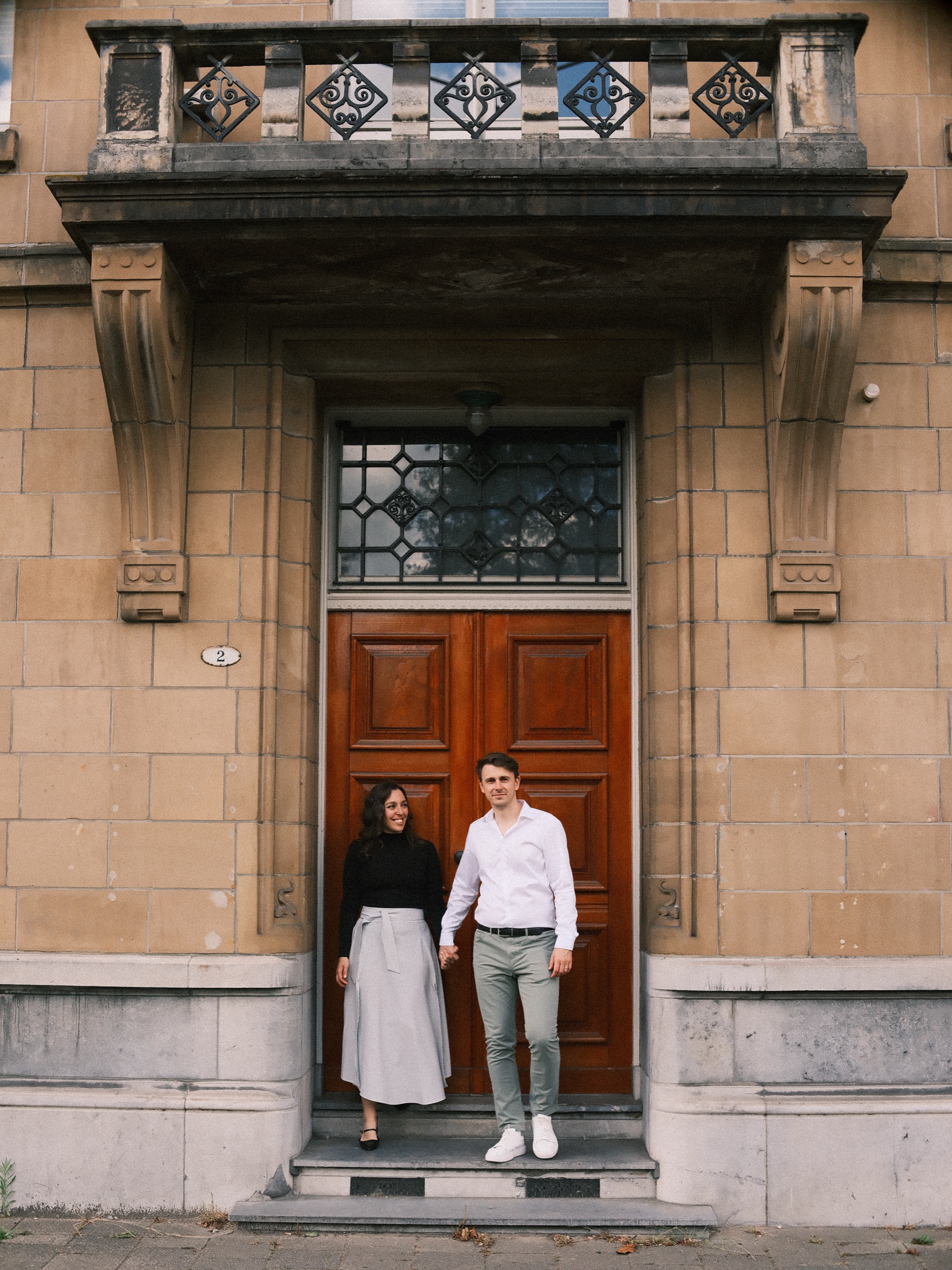 Couple stepping down from a tall red-brown door with stained glass under a stone balcony, a photo spot in the Rotterdam Scheepvaartkwartier
