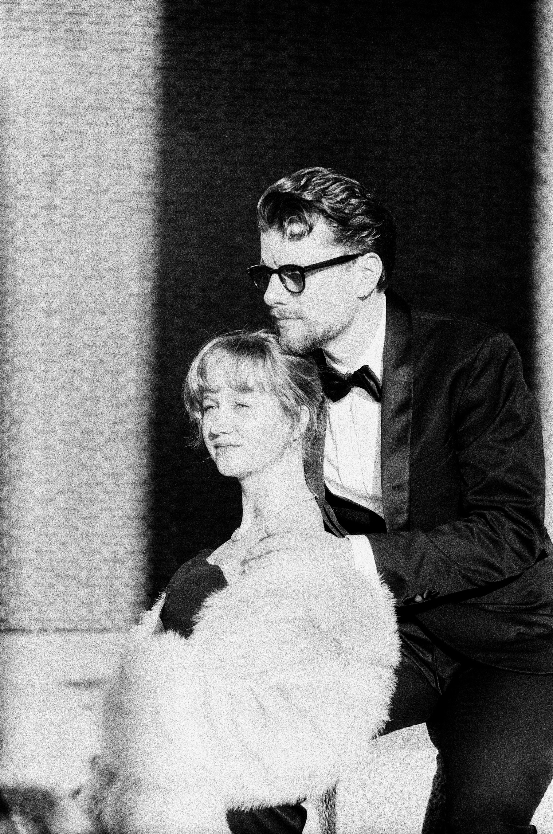 Woman with blond bangs and a pale fur coat leaning on a man in a tuxedo and bow tie, sunlight on their faces by a brick wall