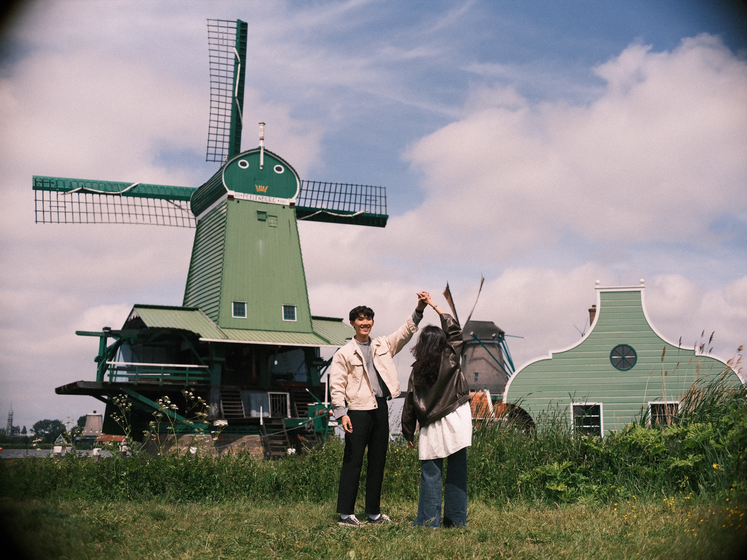He twirls her under his raised arm on the grass before De Gekroonde Poelenburg, a green mill with a gold crown on its cap, at the Zaanse Schans