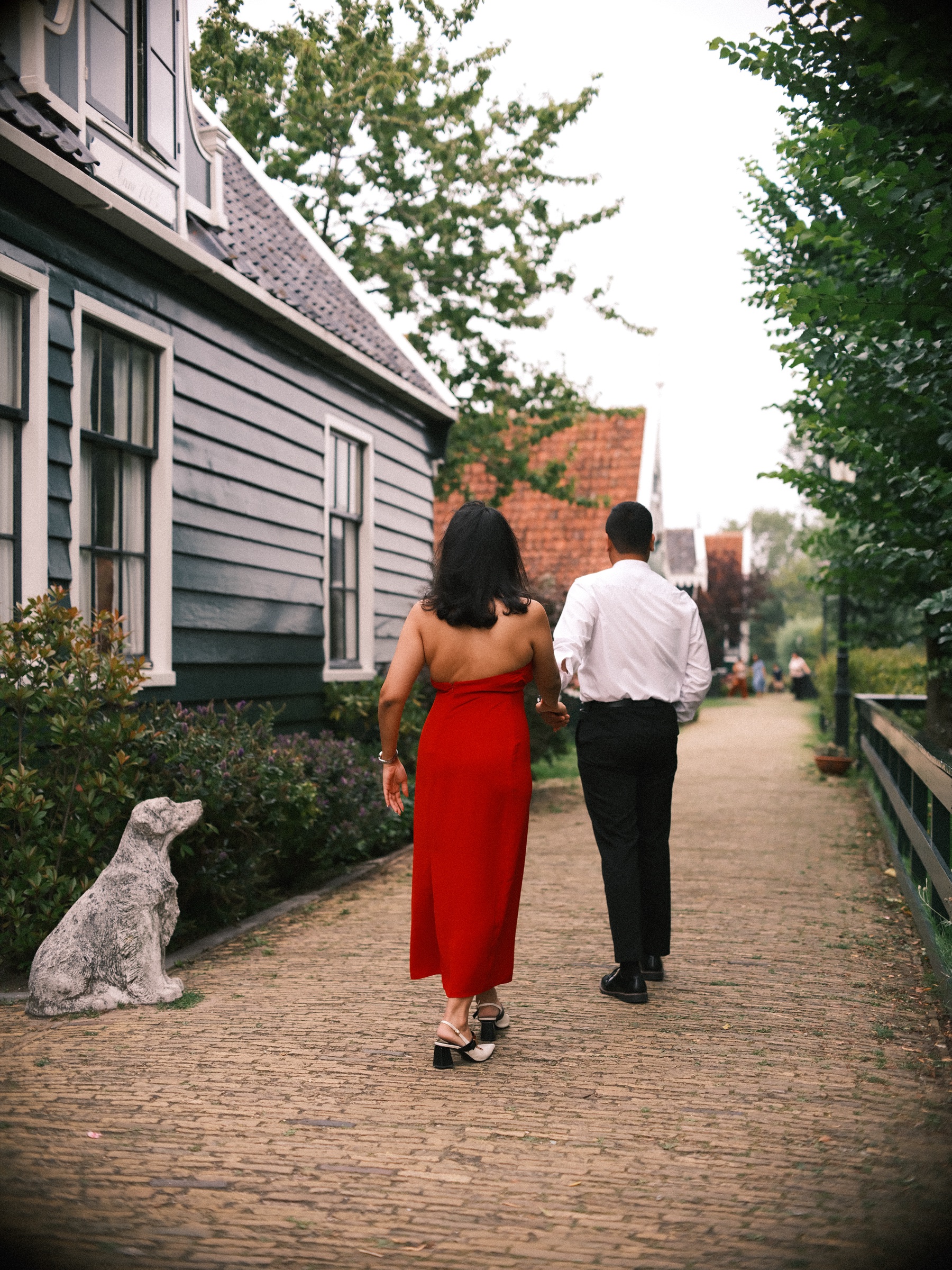 Couple walking away hand in hand down a quiet brick lane past a stone dog statue, tall trees and a dark wooden fence ahead