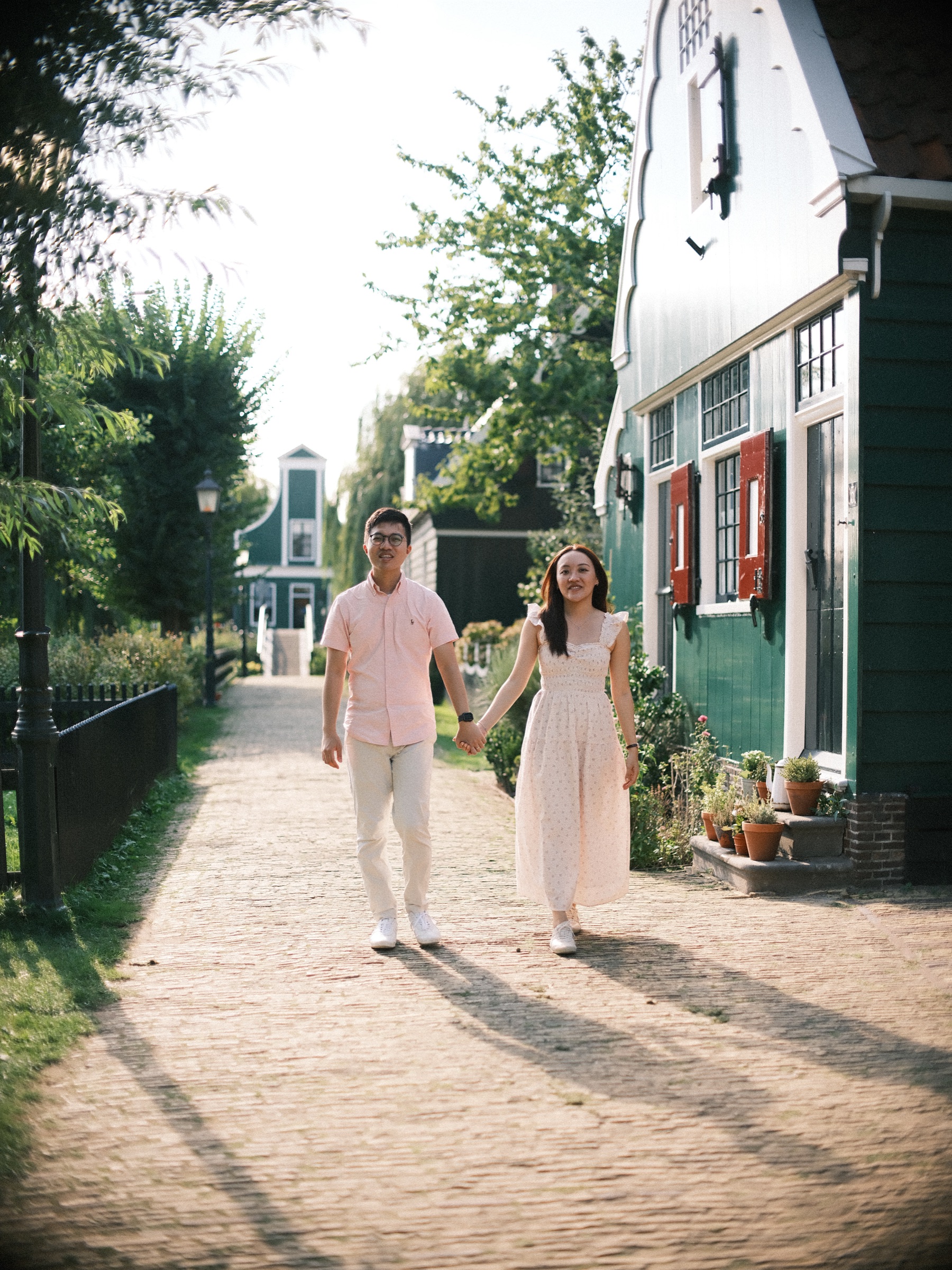 Couple in pastel summer clothes walking hand in hand down a sunlit brick path between green wooden houses, long shadows behind them