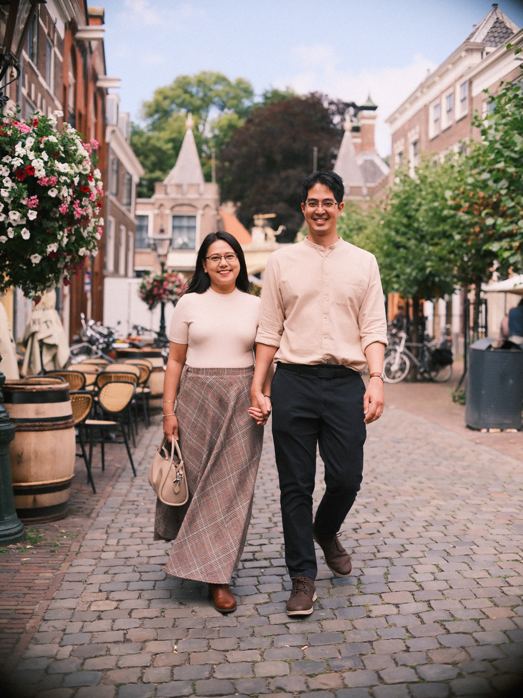 Couple walking hand in hand down a cobbled café lane in Leiden, wine barrels, terrace chairs and hanging flower baskets around them
