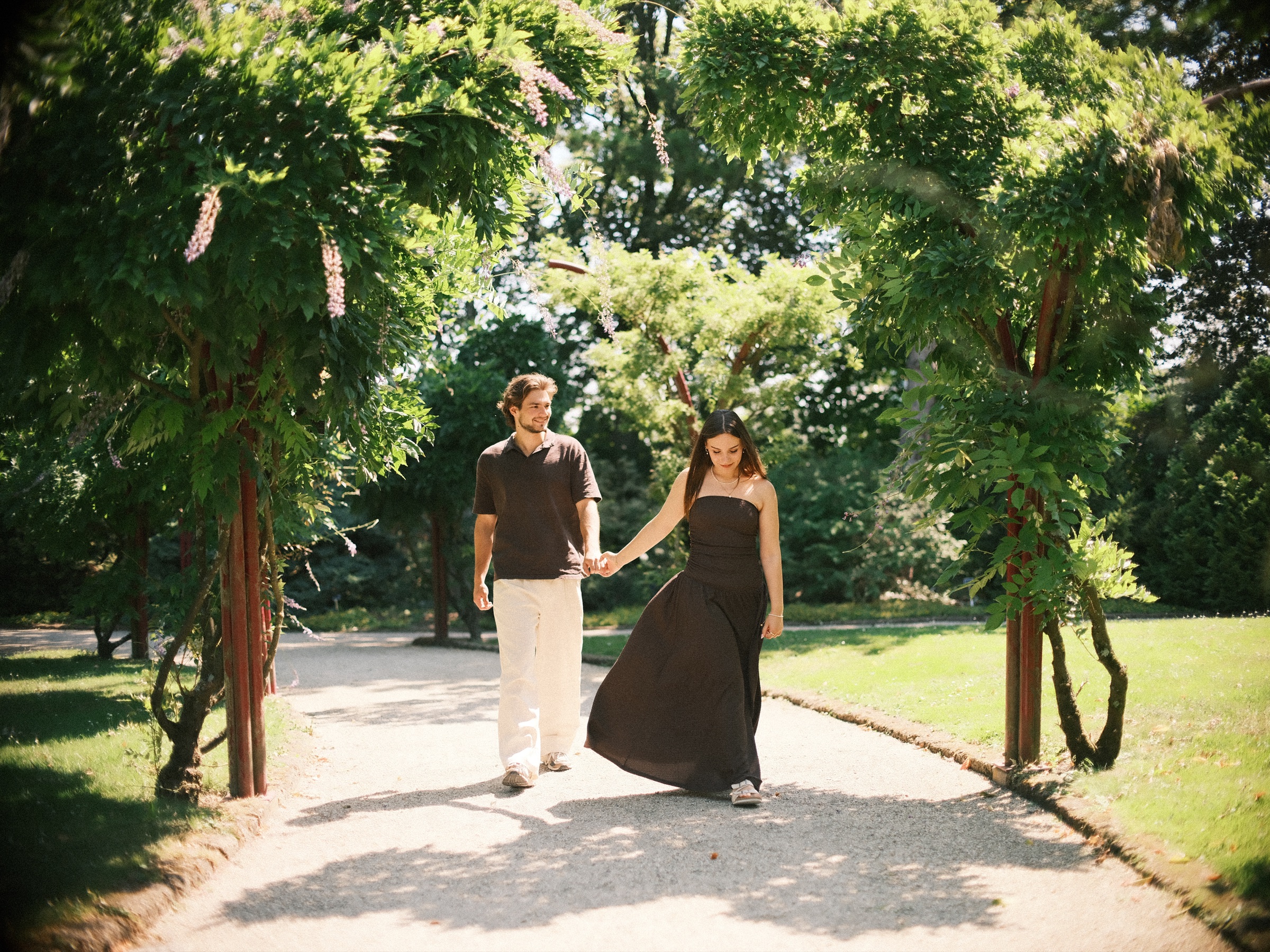 Hand in hand under a long green arch, she swings her brown maxi dress on a sunny path at Trompenburg before the proposal