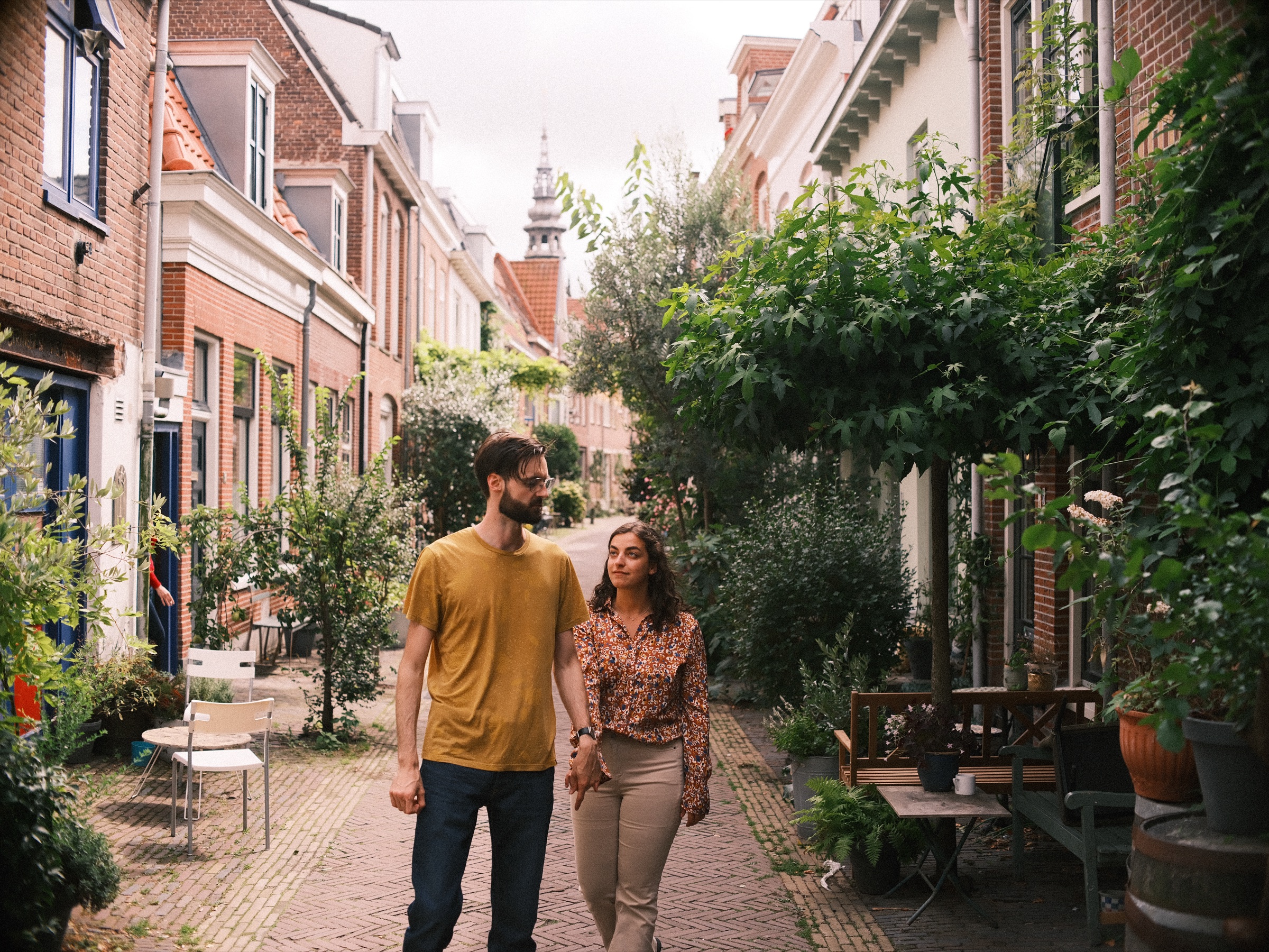 Stel loopt hand in hand door een smal straatje vol planten en bankjes bij de Vijfhoek, met een kerktoren aan het eind van de straat