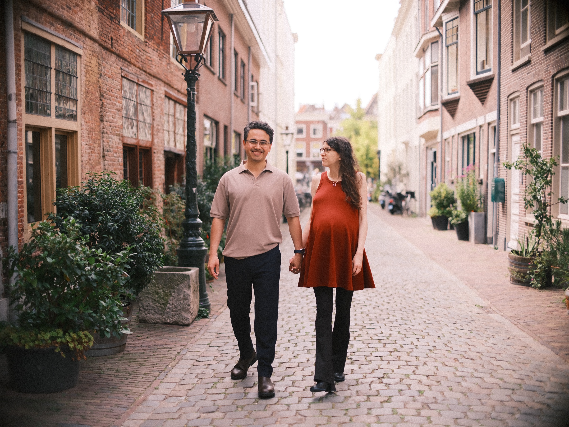 Expecting couple walking side by side down a narrow cobbled lane with lanterns and potted plants on a summer day in Leiden