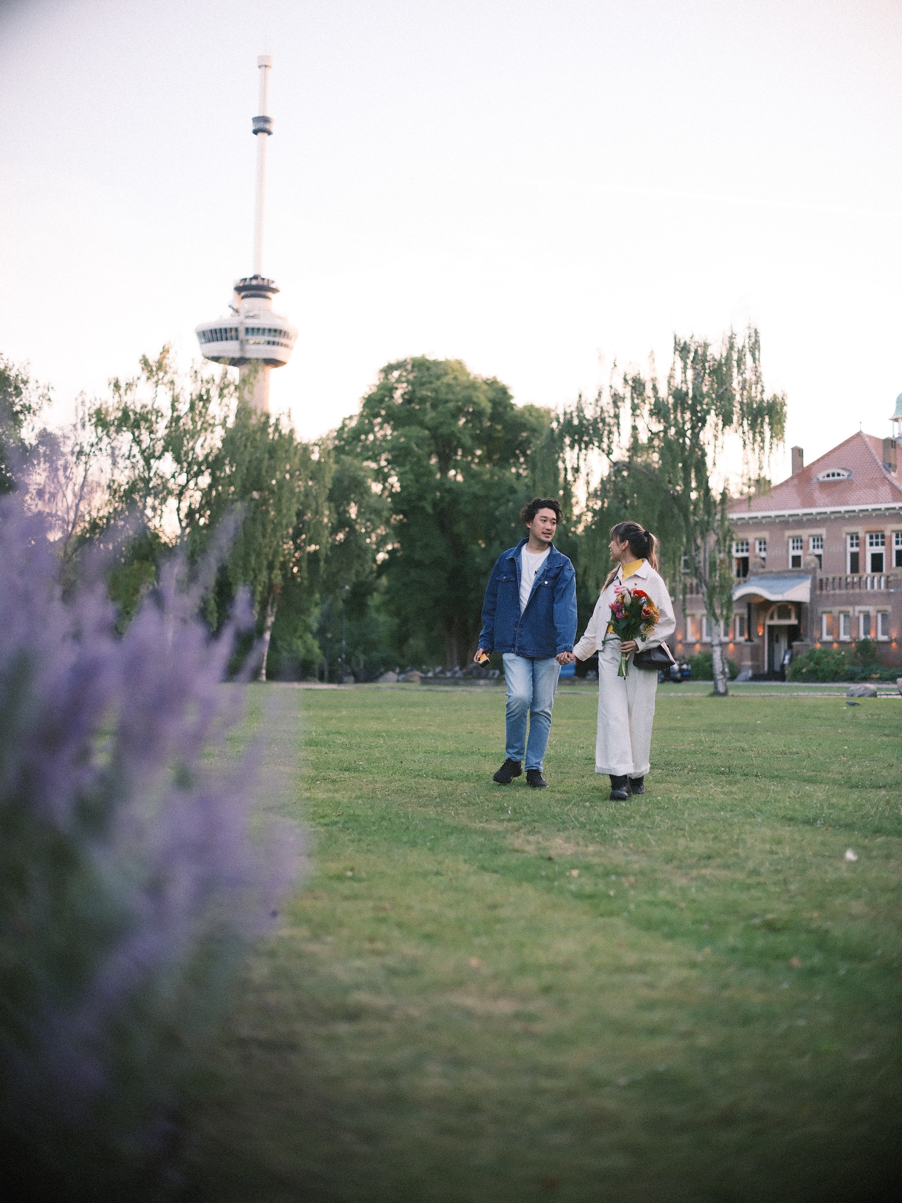 Stel loopt hand in hand over een groot grasveld, met paarse bloemen wazig vooraan en de Euromast boven een rode villa