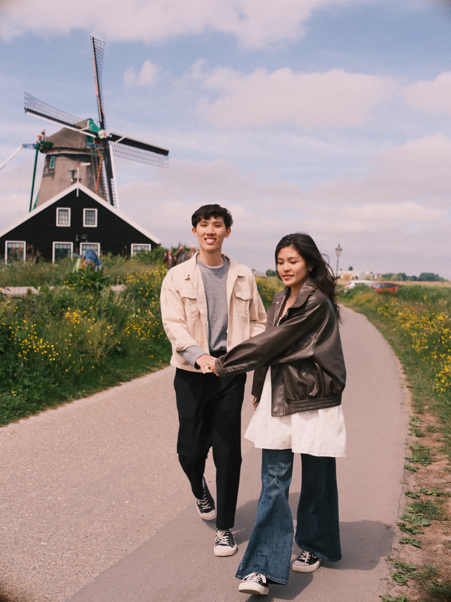 Couple walking hand in hand down a winding path toward a windmill and a dark wooden house, the last frame of their Zaanse Schans proposal