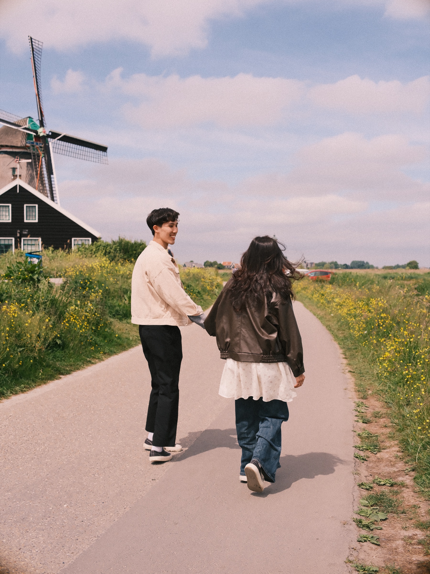 Couple walking hand in hand down a paved path lined with yellow flowers, a dark wooden mill and a black house behind them at the Zaanse Schans
