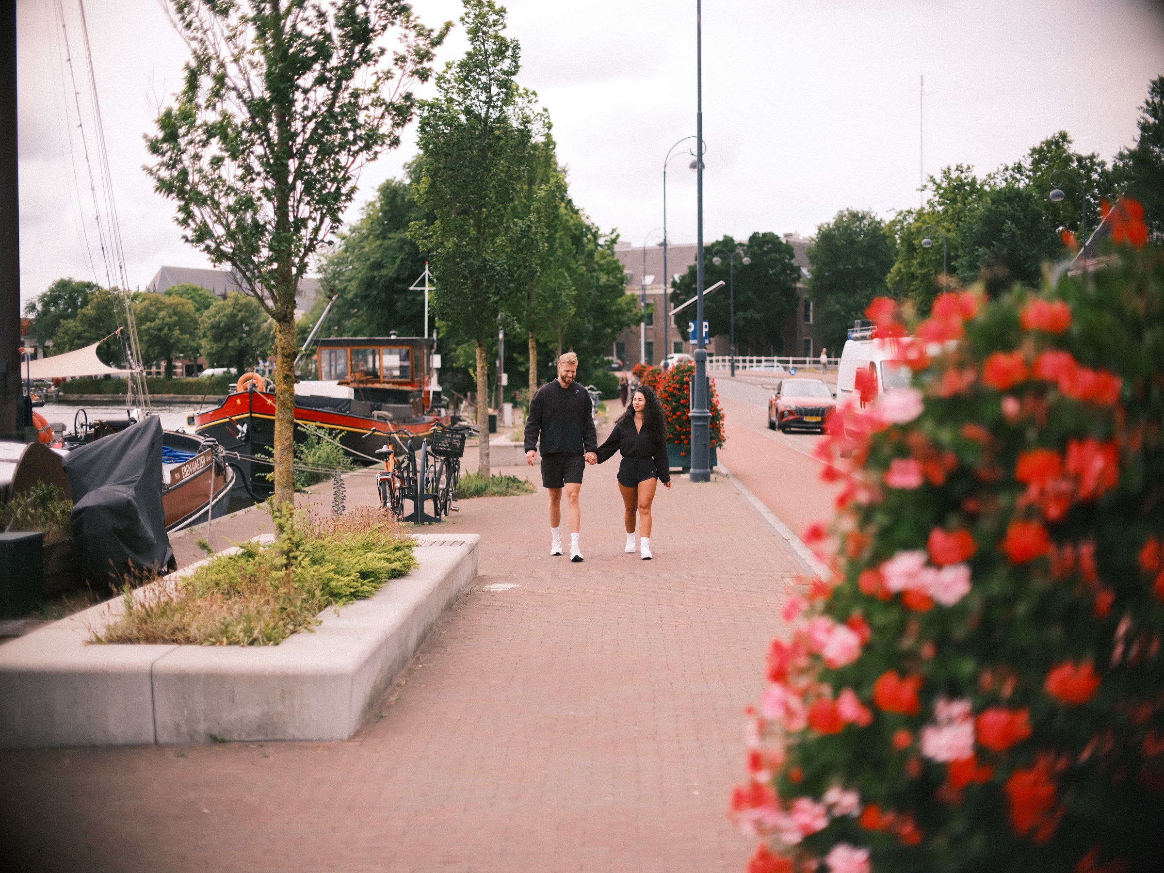 Stel loopt hand in hand over een bakstenen kade langs aangemeerde boten en lantaarnpalen, met rode en roze geraniums zacht op de voorgrond