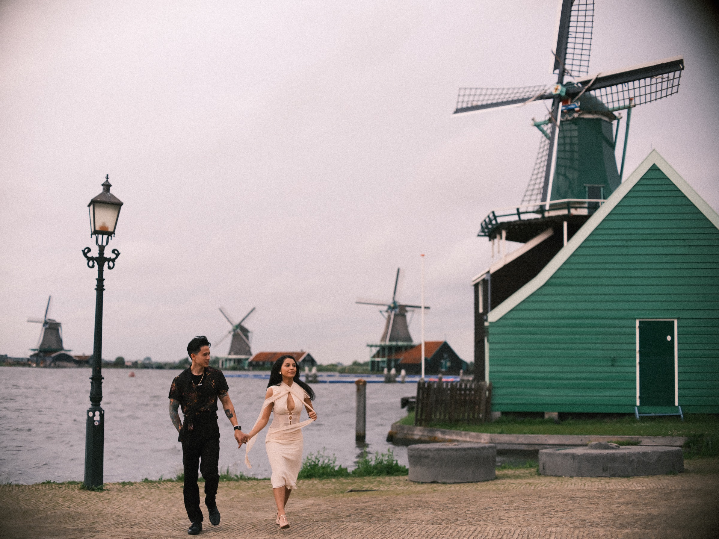 Couple walking hand in hand along the quay past an old lamppost and a green mill house, the windmills of the Zaanse Schans across the Zaan
