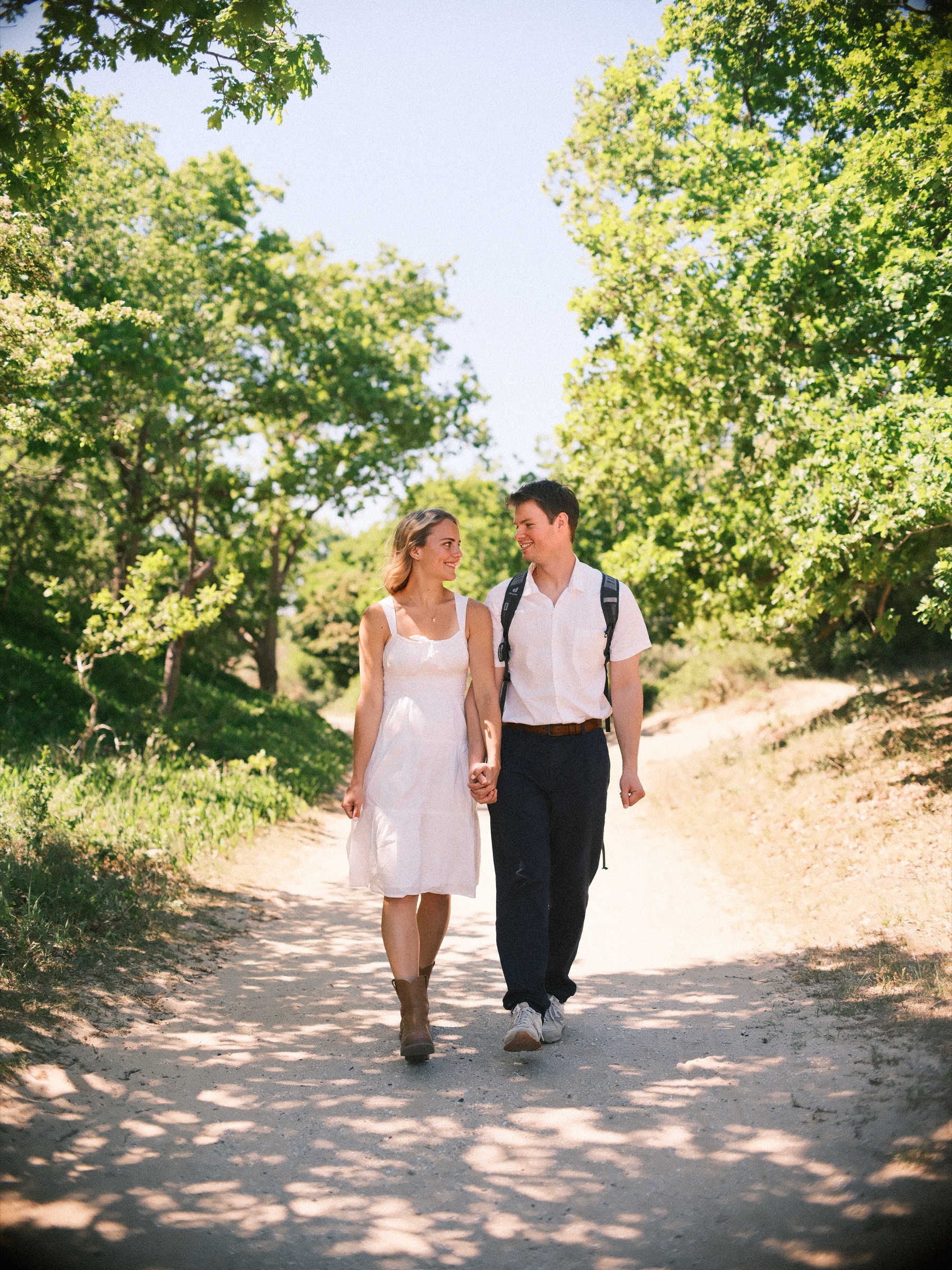 Man met rugzak en vrouw in een witte jurk lopen hand in hand over een zandpad tussen groene struiken en bomen