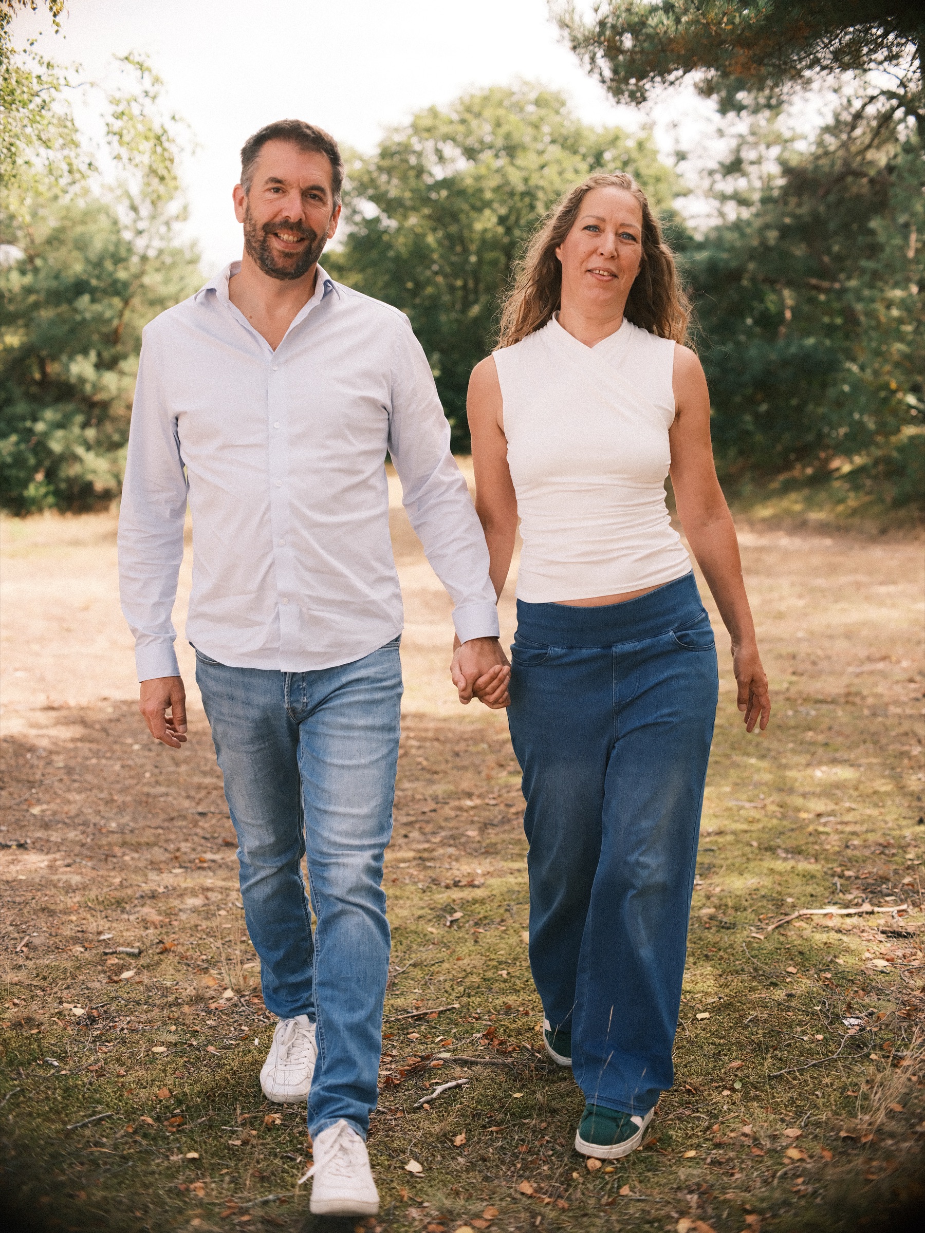 Couple walking hand in hand along a sandy path between pines and green bushes, both smiling on a sunny late summer day