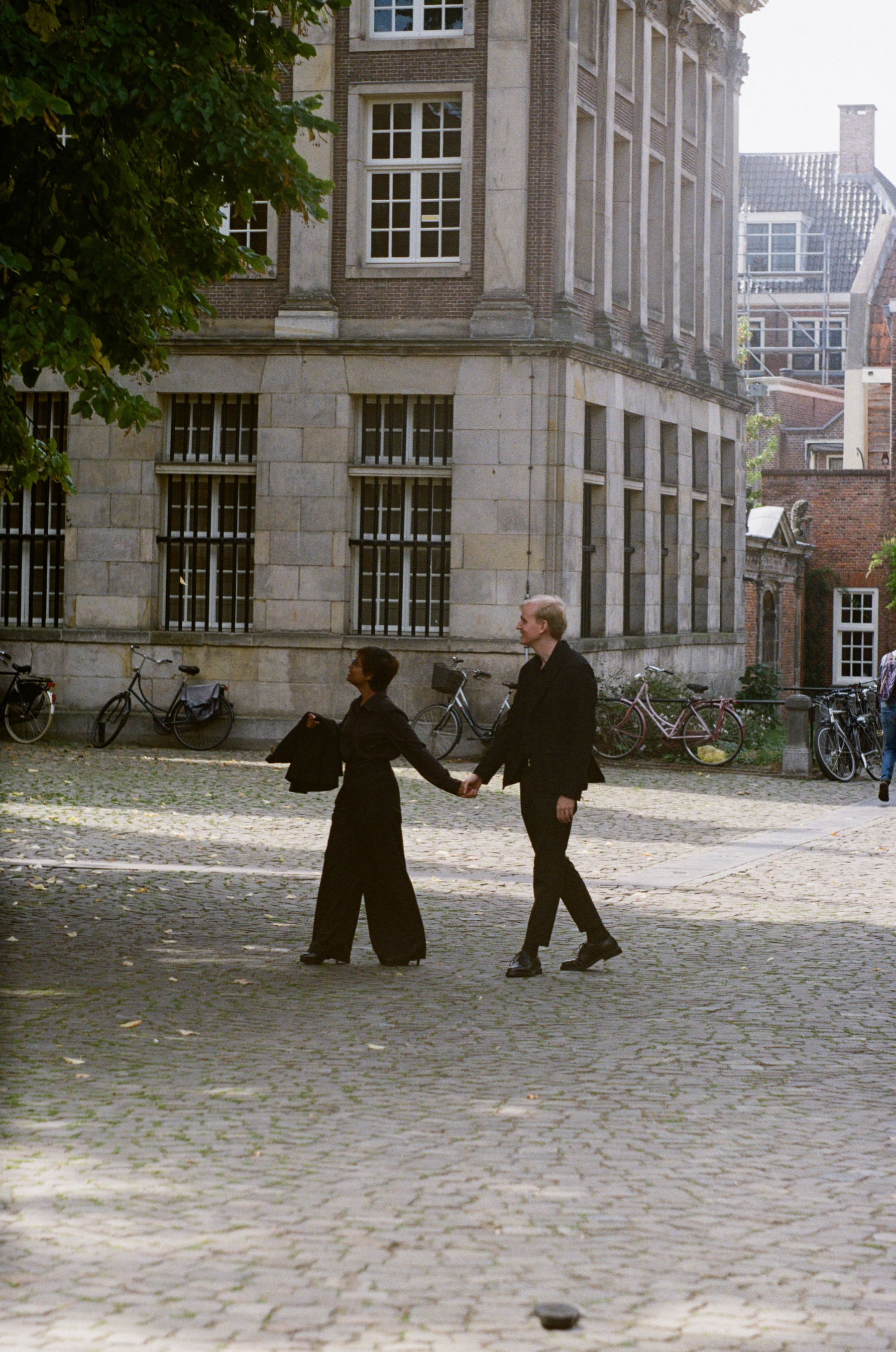 Couple walking hand in hand across a cobbled square past a grand stone building, a warm color frame on Kodak Gold film