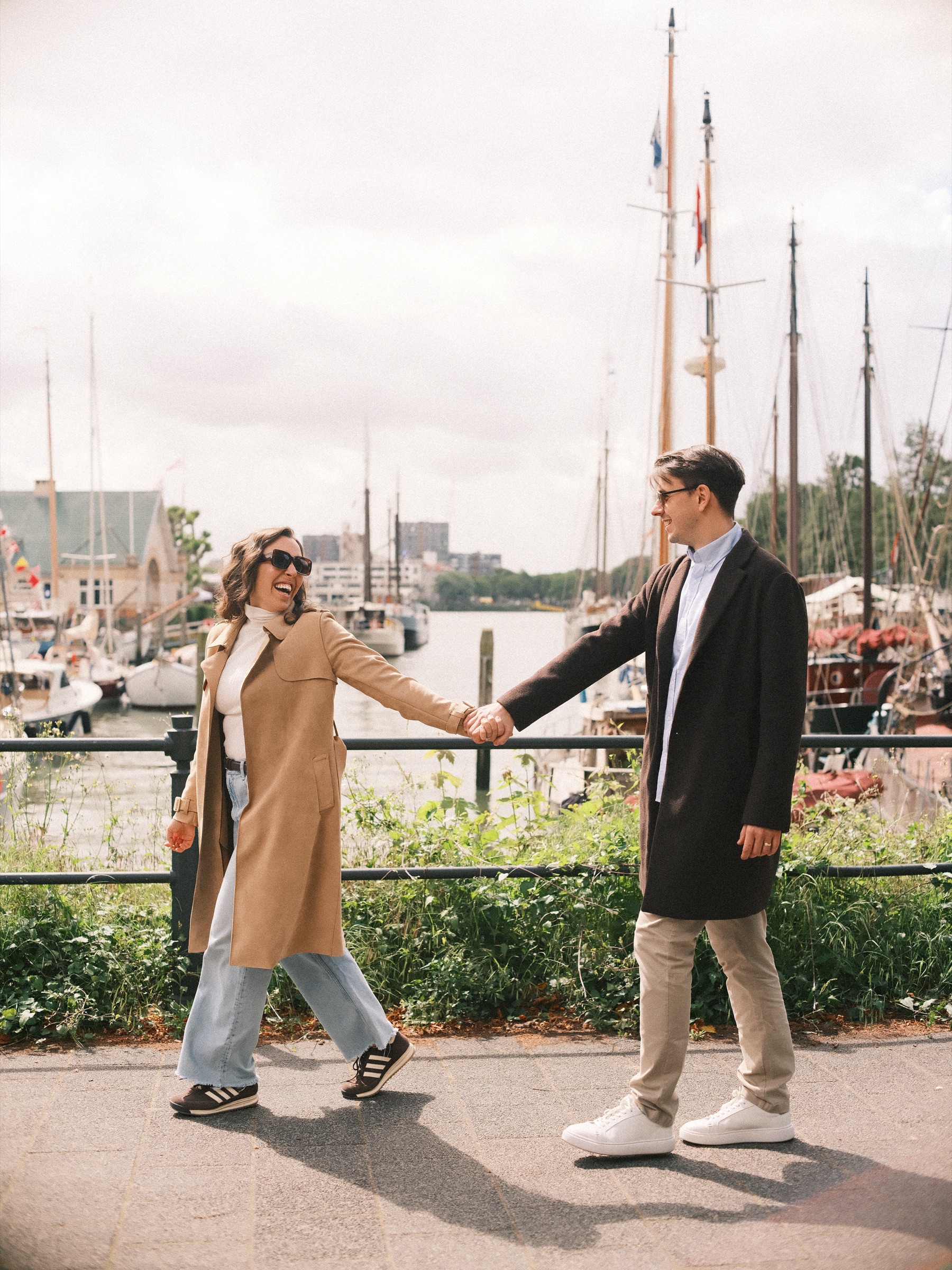Laughing woman in a camel trench coat walking hand in hand with her fiancé along the Veerhaven railing, during an engagement photo session in Rotterdam