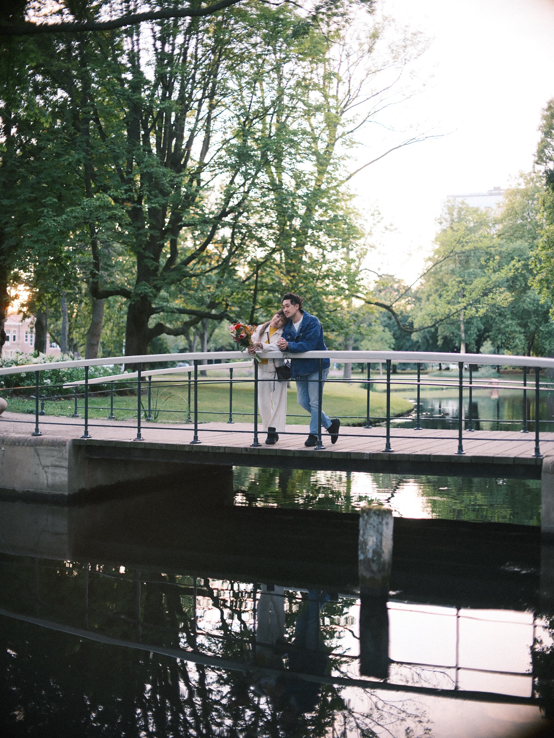 Stel leunt op de witte reling van een bruggetje in Het Park, de vijver eronder spiegelt de bomen en de brug