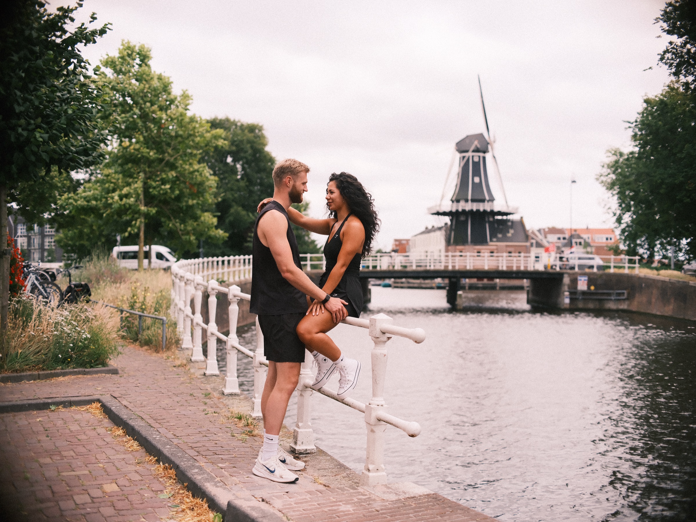 Vrouw zit op een gebogen wit hek naast haar partner, met De Adriaan en een lage brug aan de overkant van het Spaarne in Haarlem