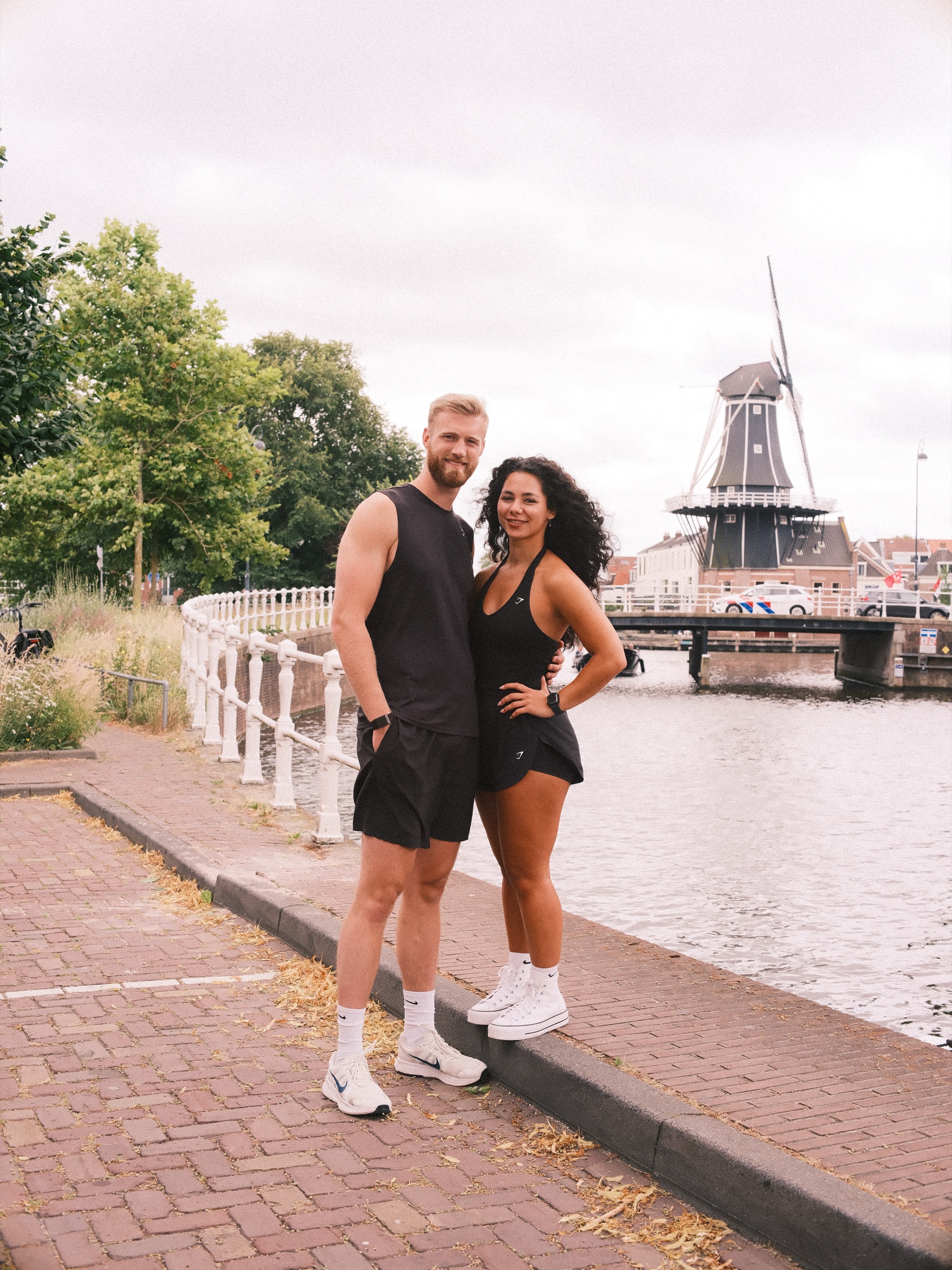 Stel in mouwloze zwarte kleren bij een gebogen wit hek, met molen De Adriaan en een brug op de achtergrond