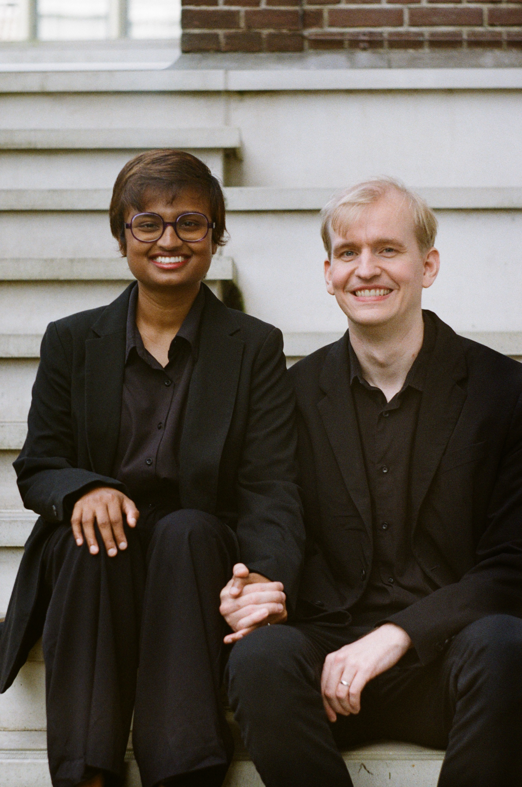 Smiling couple in black sitting close on white stone steps, holding hands, a sharp color portrait on Kodak Gold 200 film