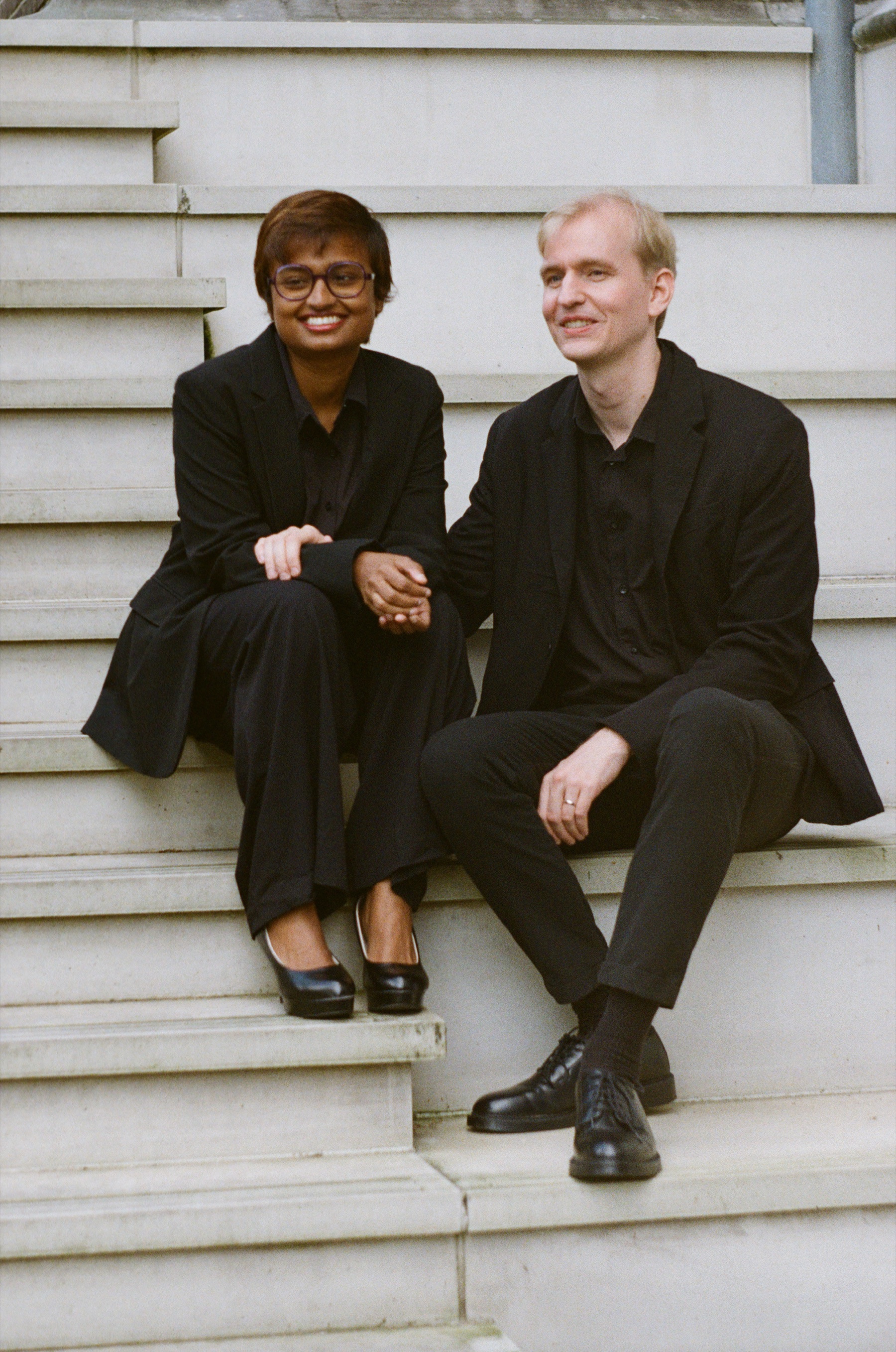 Couple in black sitting hand in hand on white stone steps in the sun, both smiling, a warm color frame on Kodak Gold 200 film