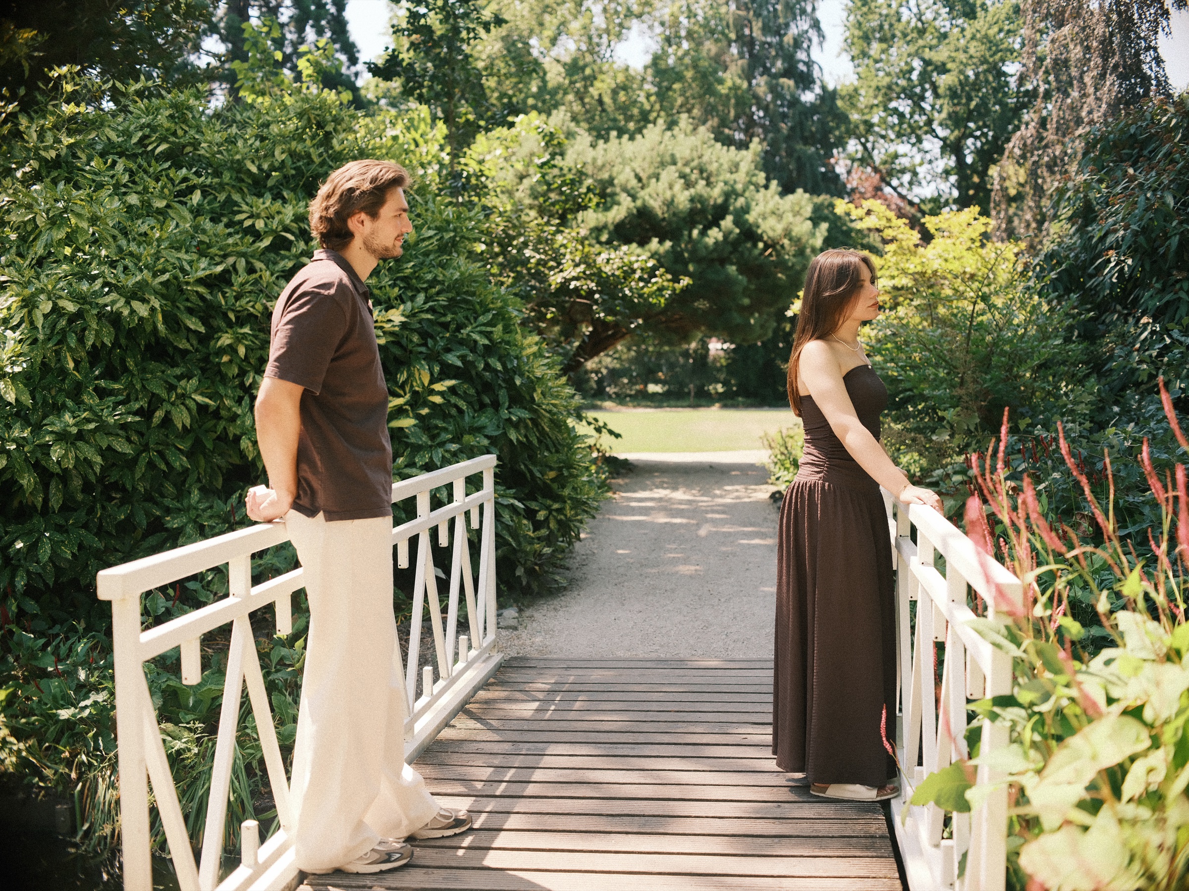 On the white bridge in the Trompenburg gardens, he leans on the railing and smiles at her as she stands among red flowers
