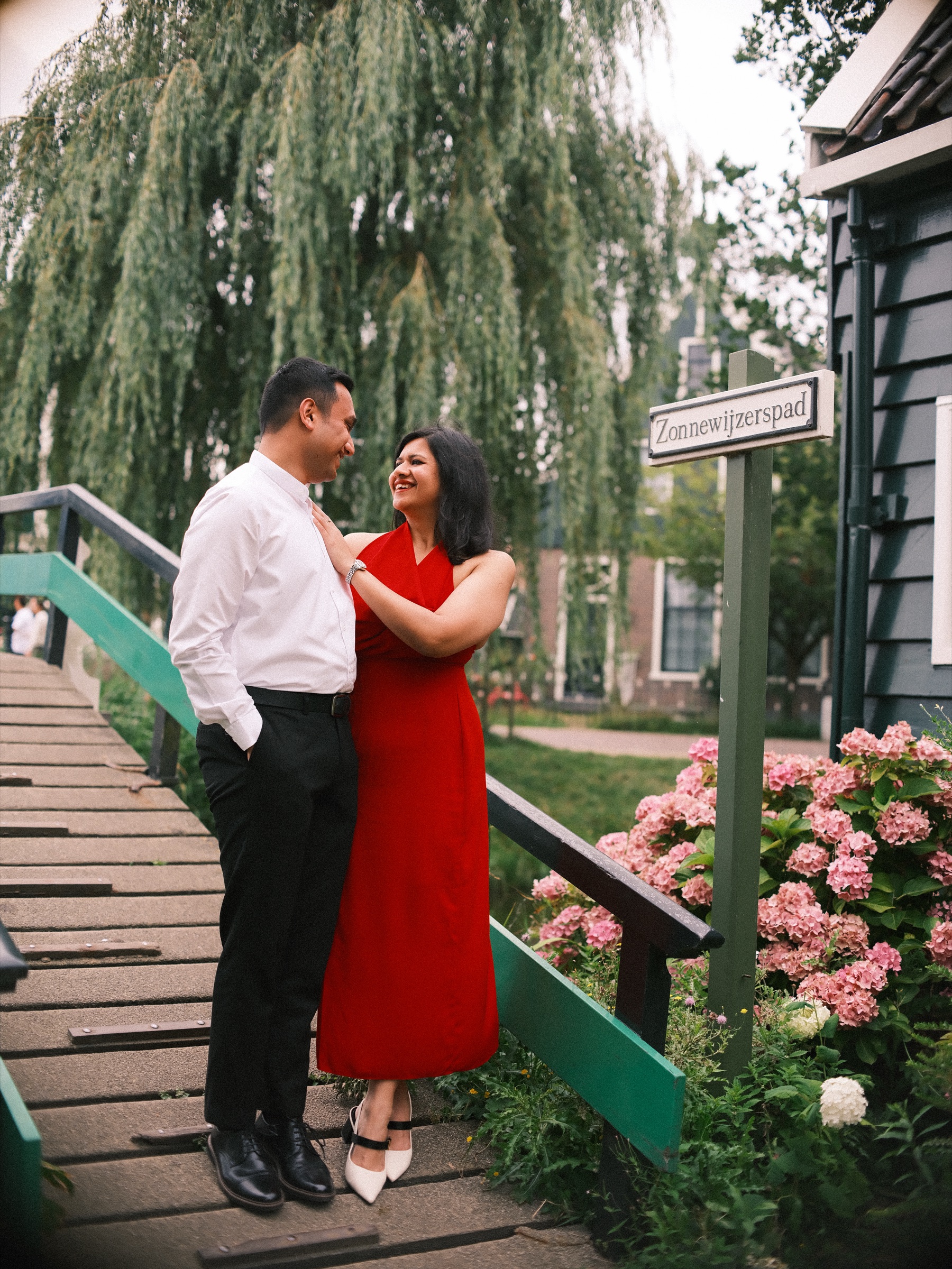 Couple on a small wooden bridge with green railings under a weeping willow, pink hydrangeas and a Zonnewijzerspad street sign beside them