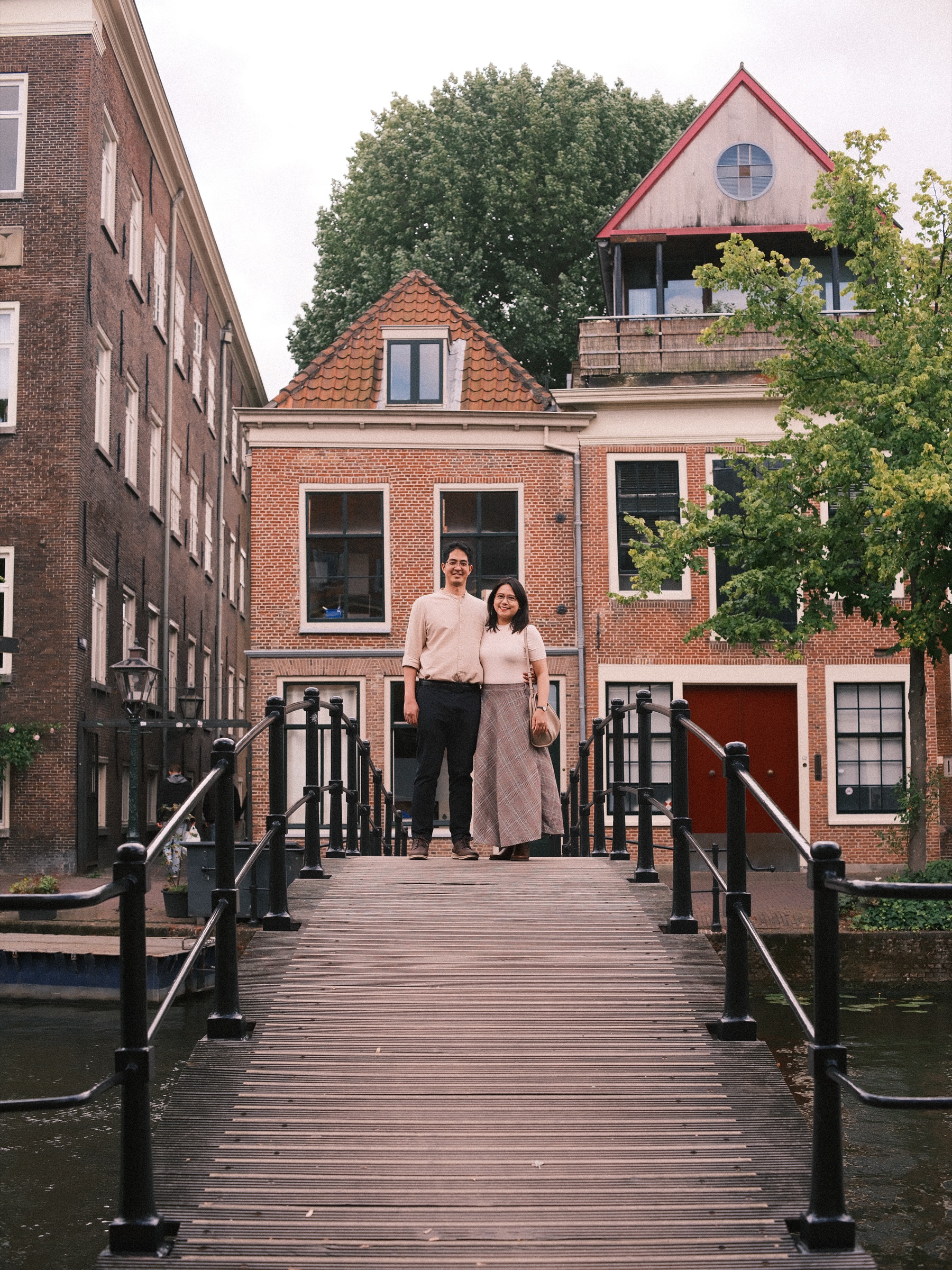 Couple at the far end of a narrow wooden footbridge with black railings, a brick house with a red front door behind them