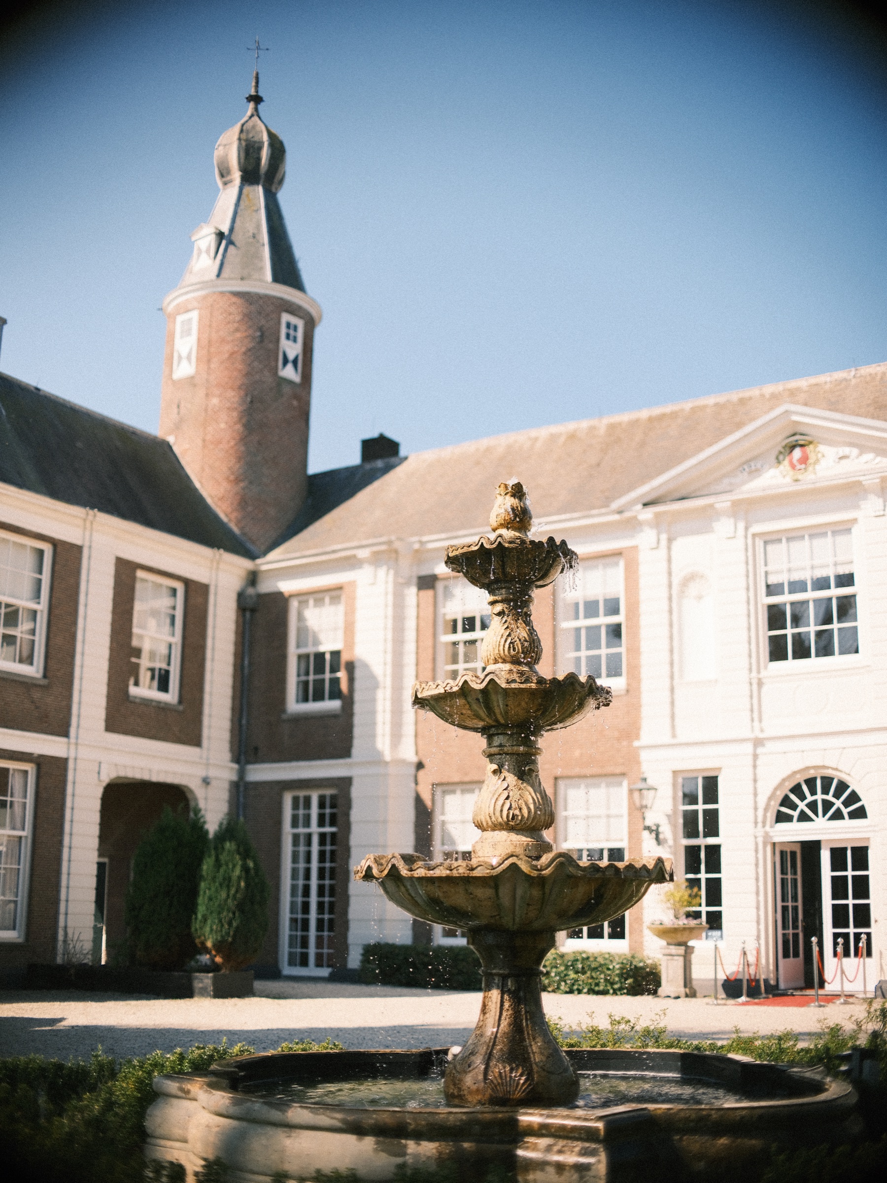 Een stenen fontein met drie schalen op de binnenplaats, terwijl de rode bakstenen toren boven de witte gevel uitsteekt