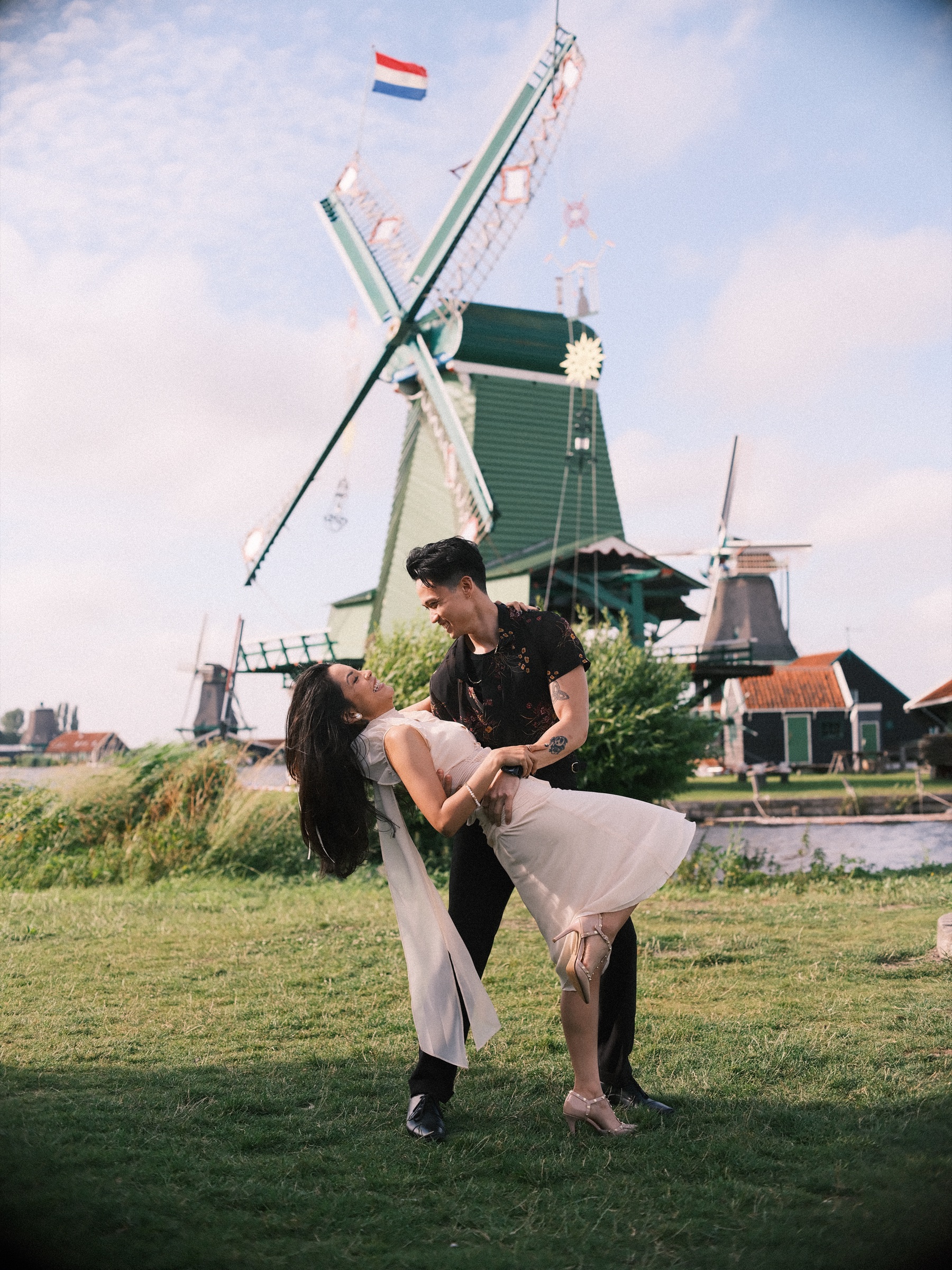 Man in a black floral shirt dipping his partner in an ivory dress under a decorated windmill with a Dutch flag at the Zaanse Schans