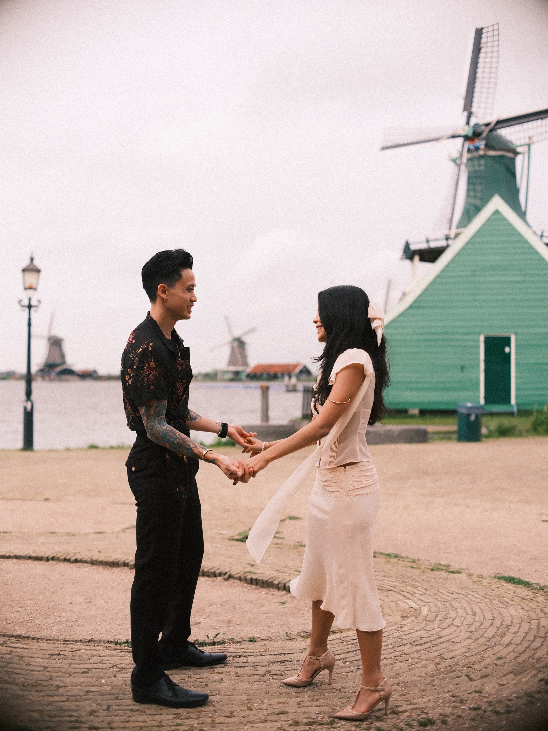 Man with a tattooed arm holding both her hands in a dance hold on a cobbled quay, a green wooden house and a lamppost behind them