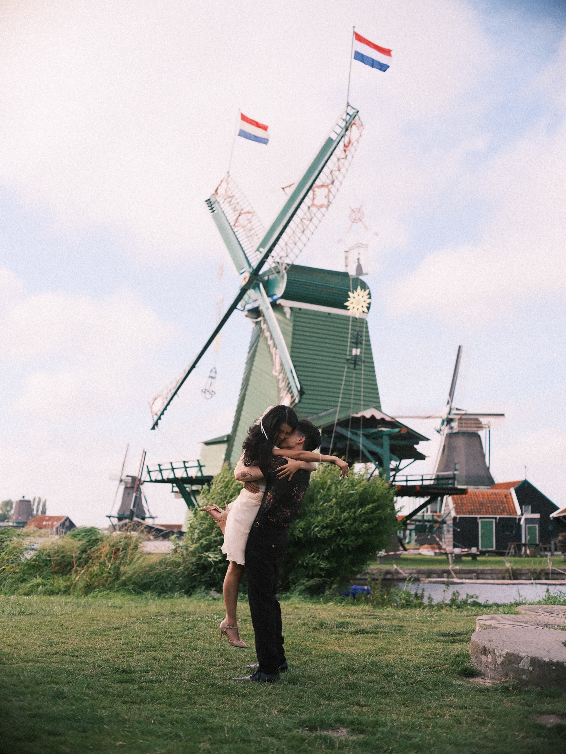Man lifting his partner high in a dance on the grass, a decorated green windmill with a Dutch flag towering above them