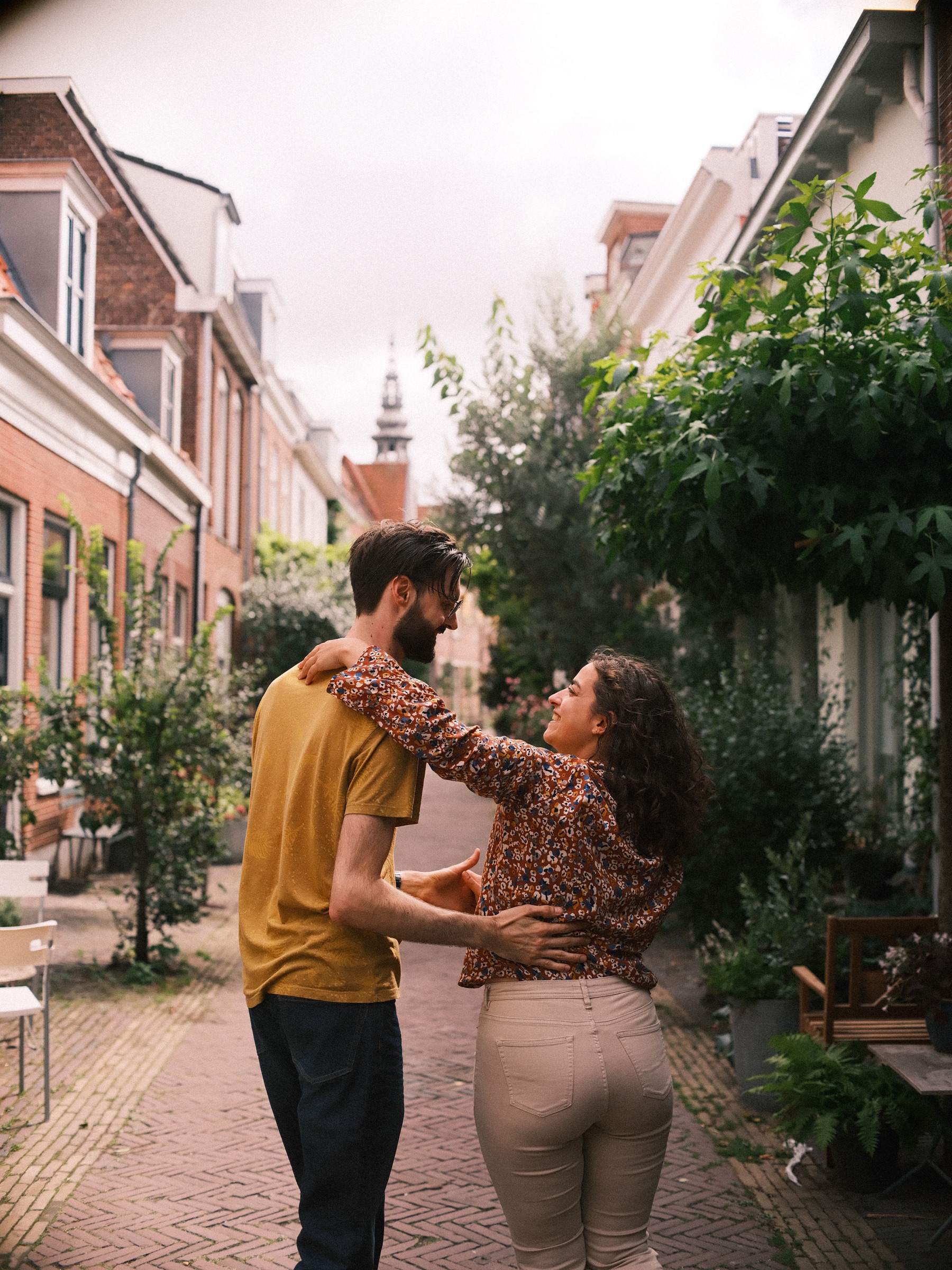 Lachende vrouw in een bloemetjesblouse danst dicht tegen haar vriend aan, met groene planten en rode bakstenen huizen rondom
