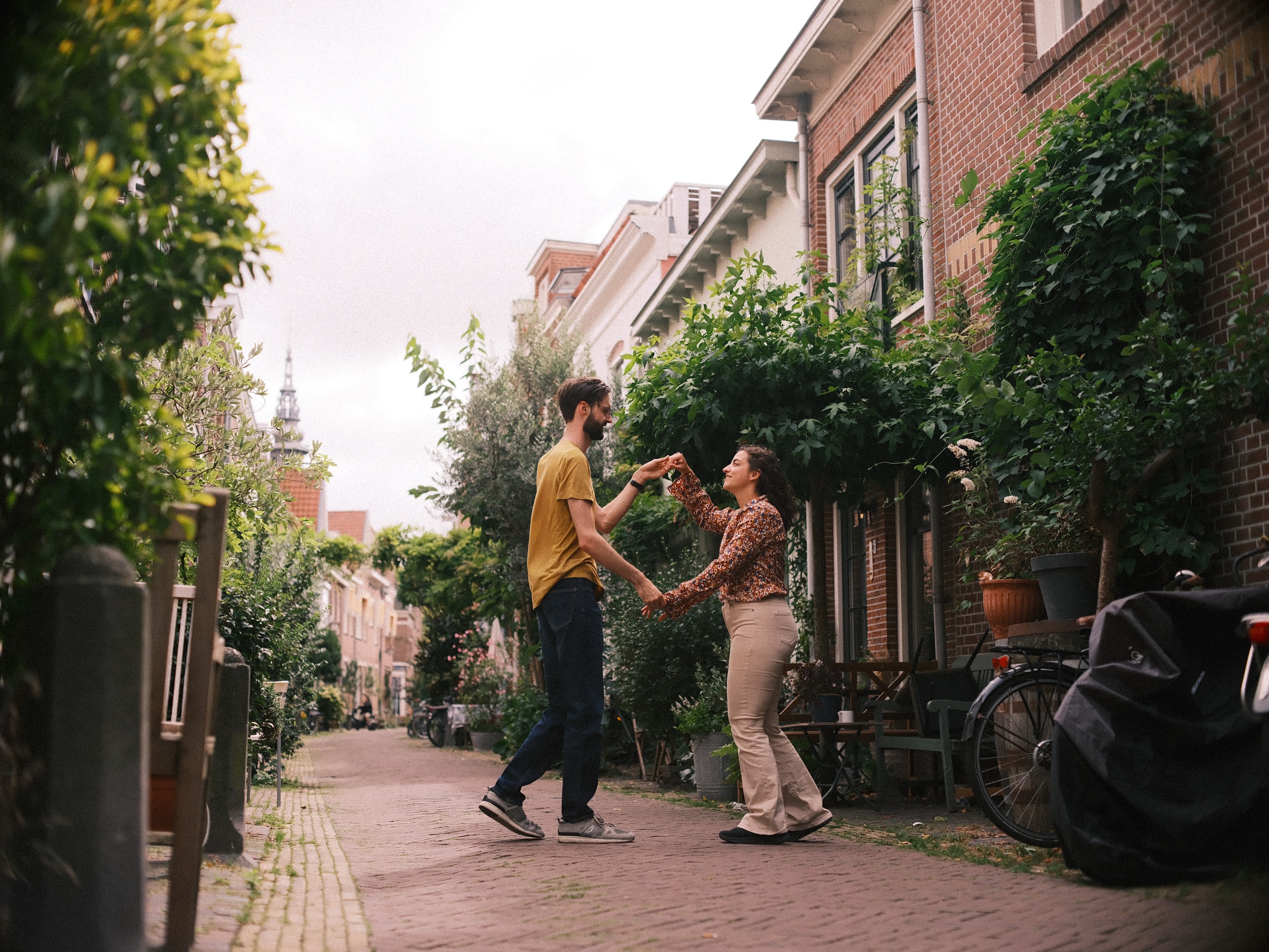 Stel danst in een smal bakstenen straatje met planten, bankjes en fietsen, met een kerktoren aan het eind van het zicht