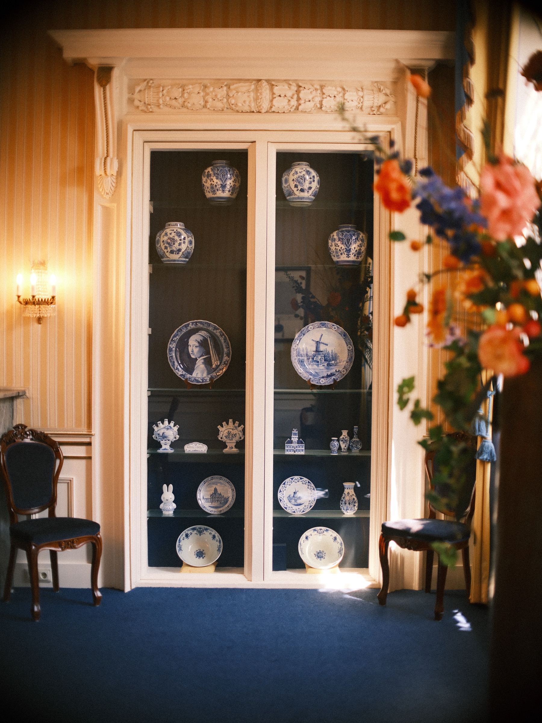 Borden van Delfts blauw in een hoge glazen kast, op één ervan het Meisje met de parel, naast oranje bloemen en een blauwe loper