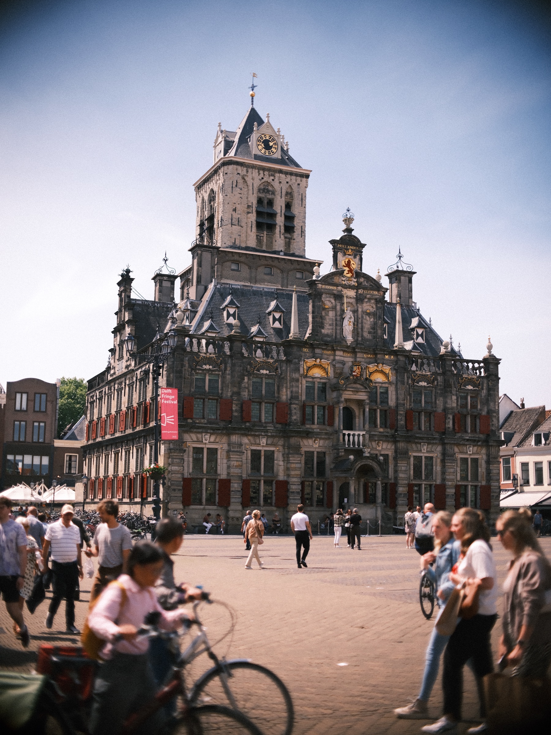 De rijk versierde gevel van het stadhuis van Delft met rode luiken en vlaggen, met fietsers en voorbijgangers op de zonnige Markt