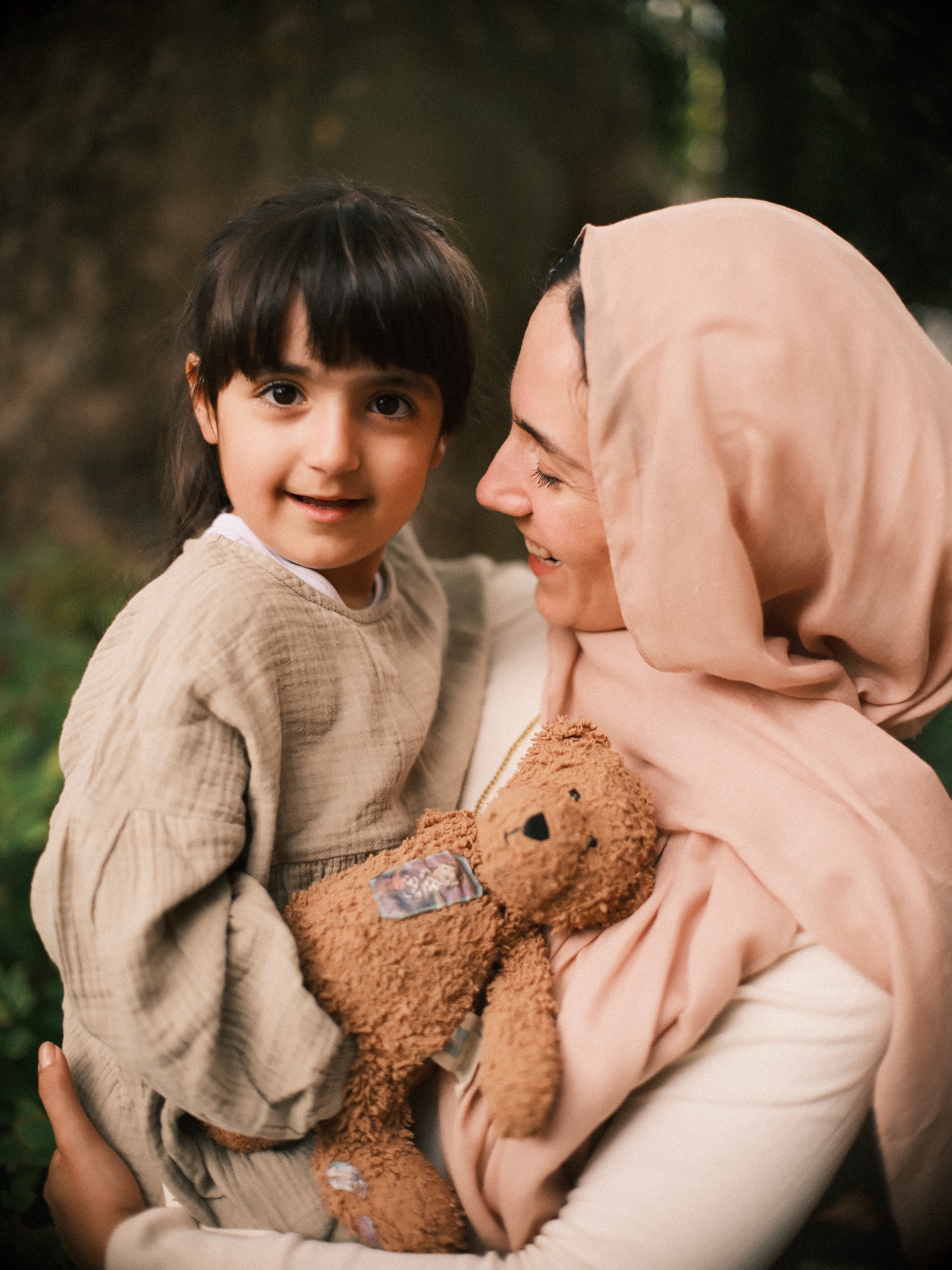 Mother in a pink headscarf holding her daughter and a brown teddy bear in a green garden in Delft, a moment from a digital photoshoot