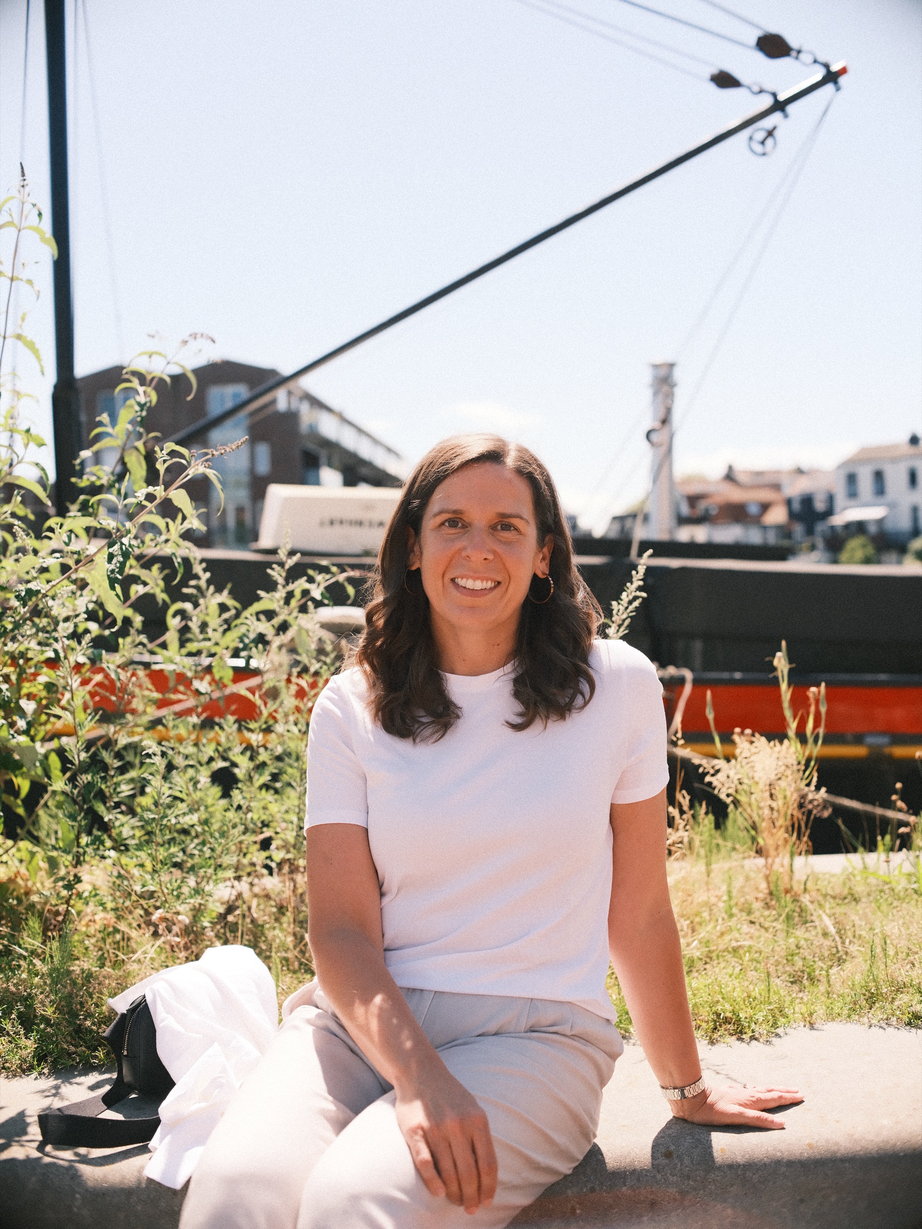 Smiling woman in a white T-shirt sitting on a low concrete edge by the Spaarne in Haarlem, an old barge behind her, from a digital photoshoot