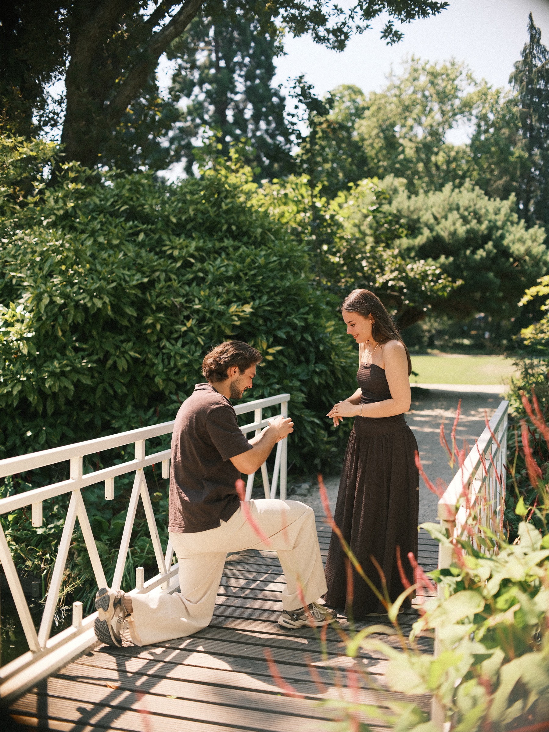 Man on one knee holding up a ring on a white wooden bridge as she smiles down at him, a digital proposal photoshoot in the Trompenburg gardens