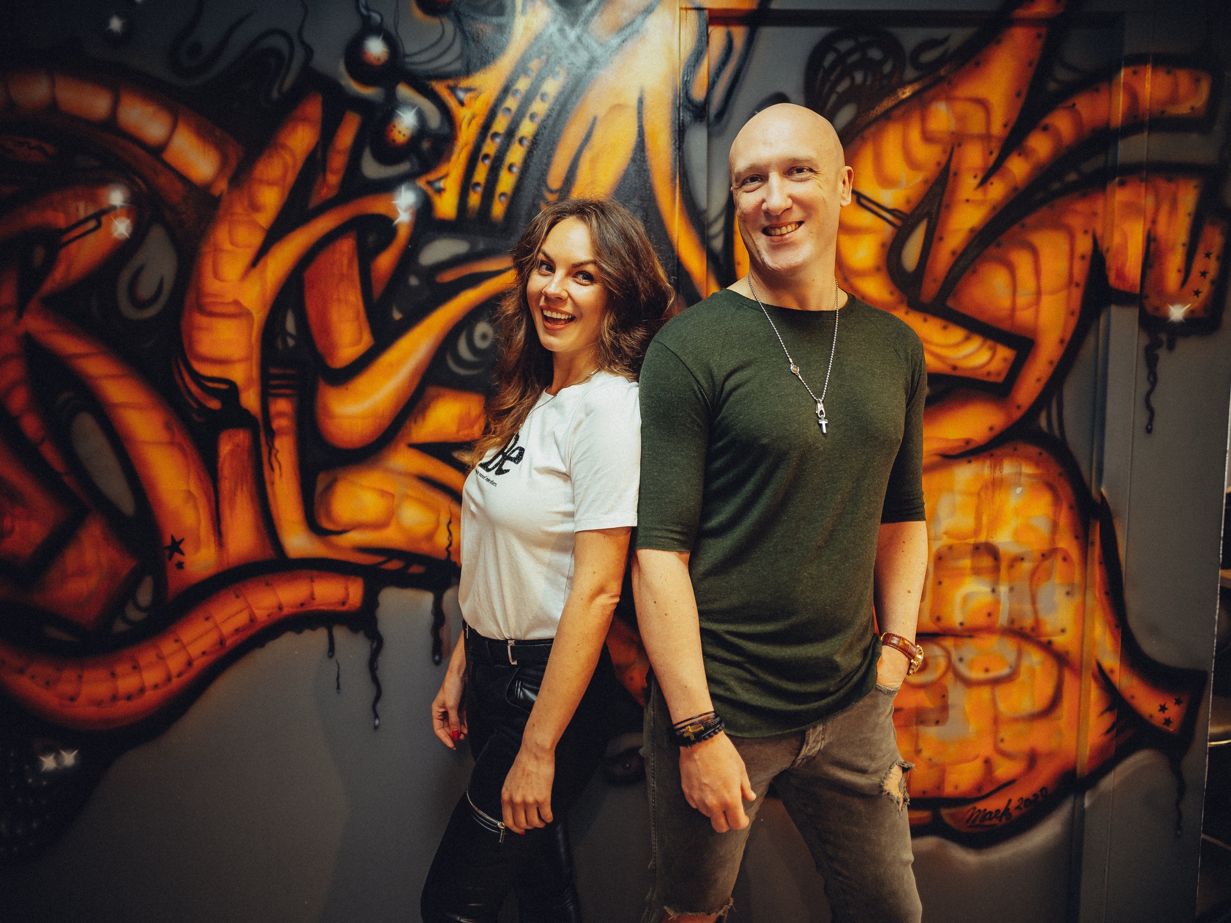 Eigenaar in een wit shirt en een man in een groen shirt lachen samen voor de oranje graffitimuur van de studio