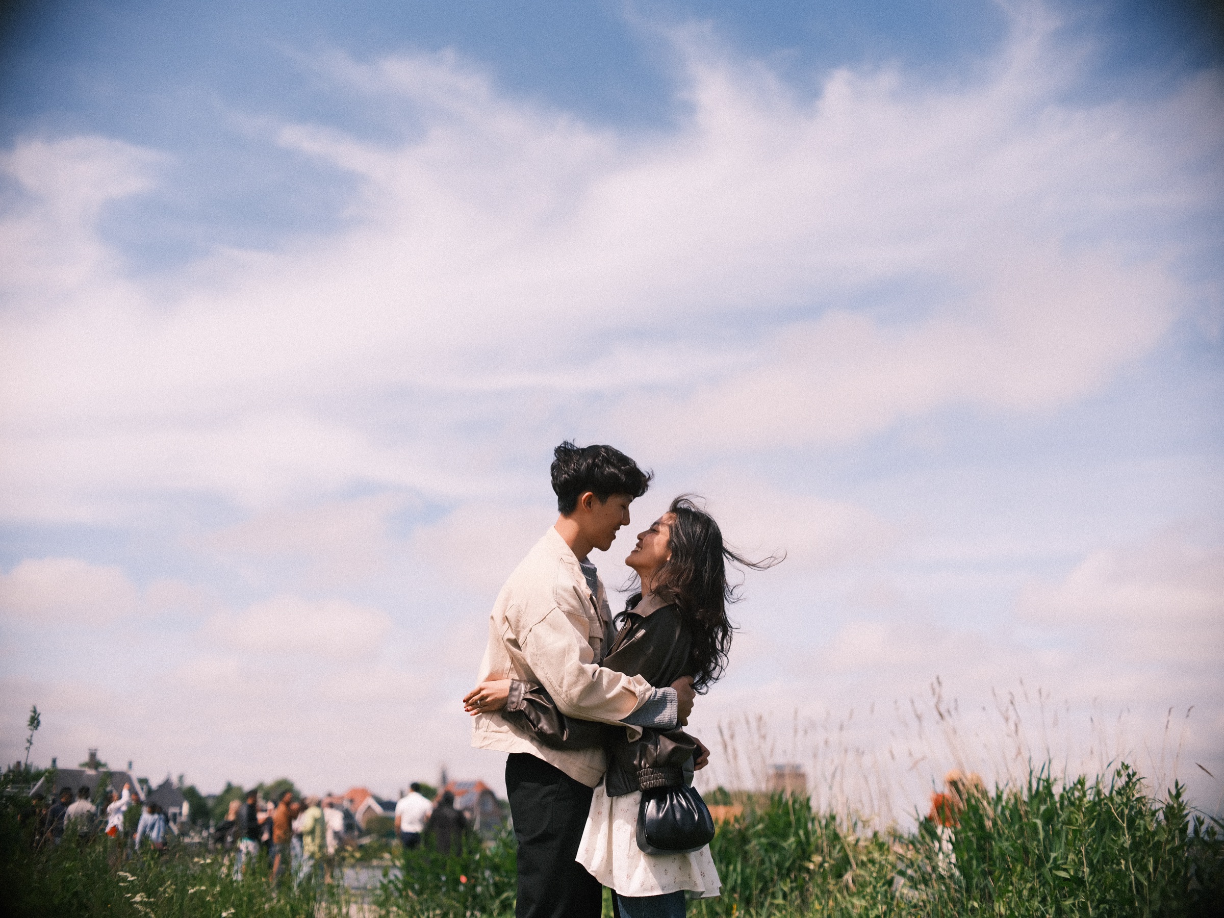 Couple in a tight embrace right after the proposal at the Zaanse Schans, white clouds streaking across a blue sky above the grass