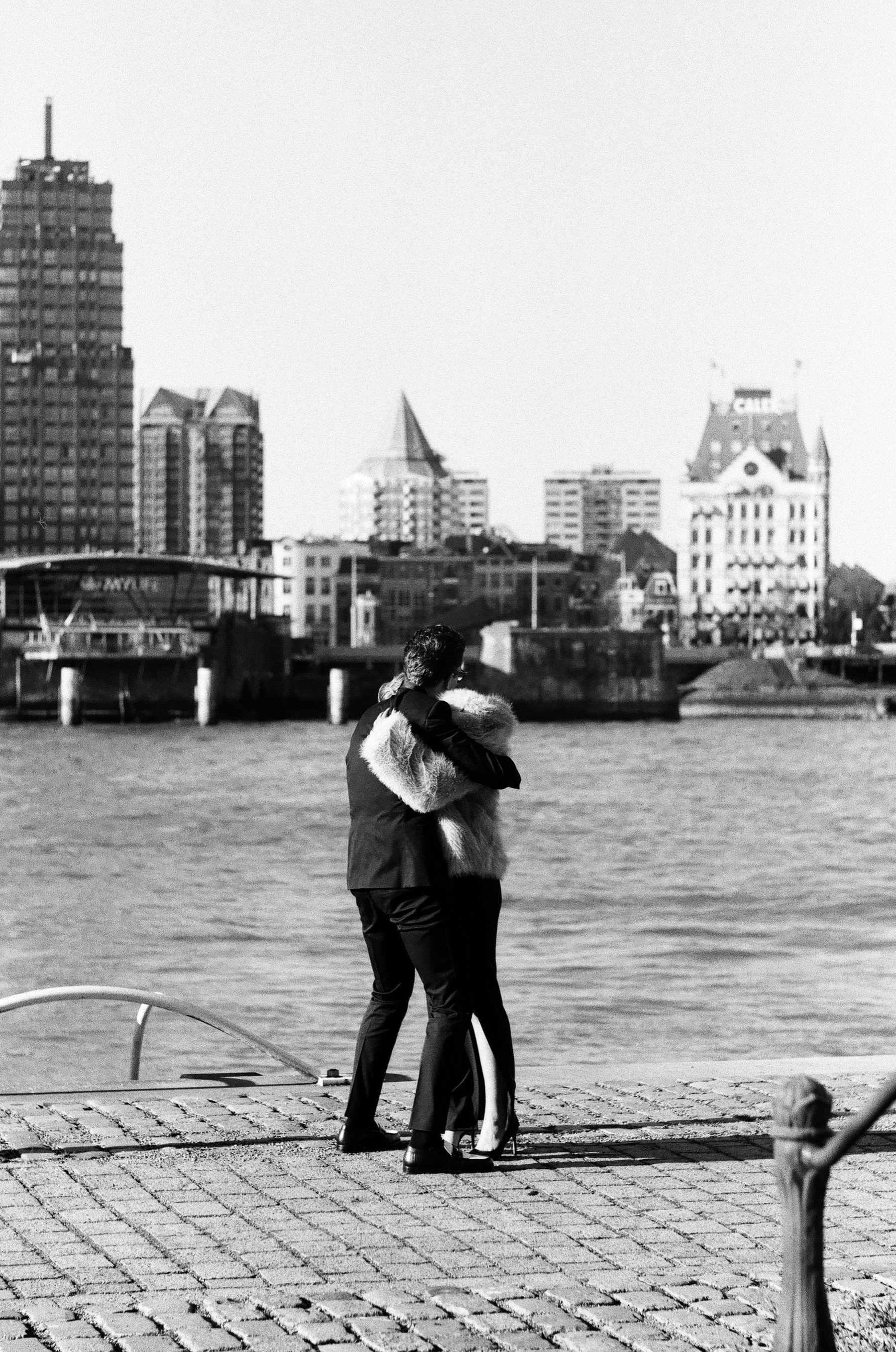 Long embrace on the cobbled quay by the Maas, towers and Het Witte Huis across the water, on black and white film in Rotterdam