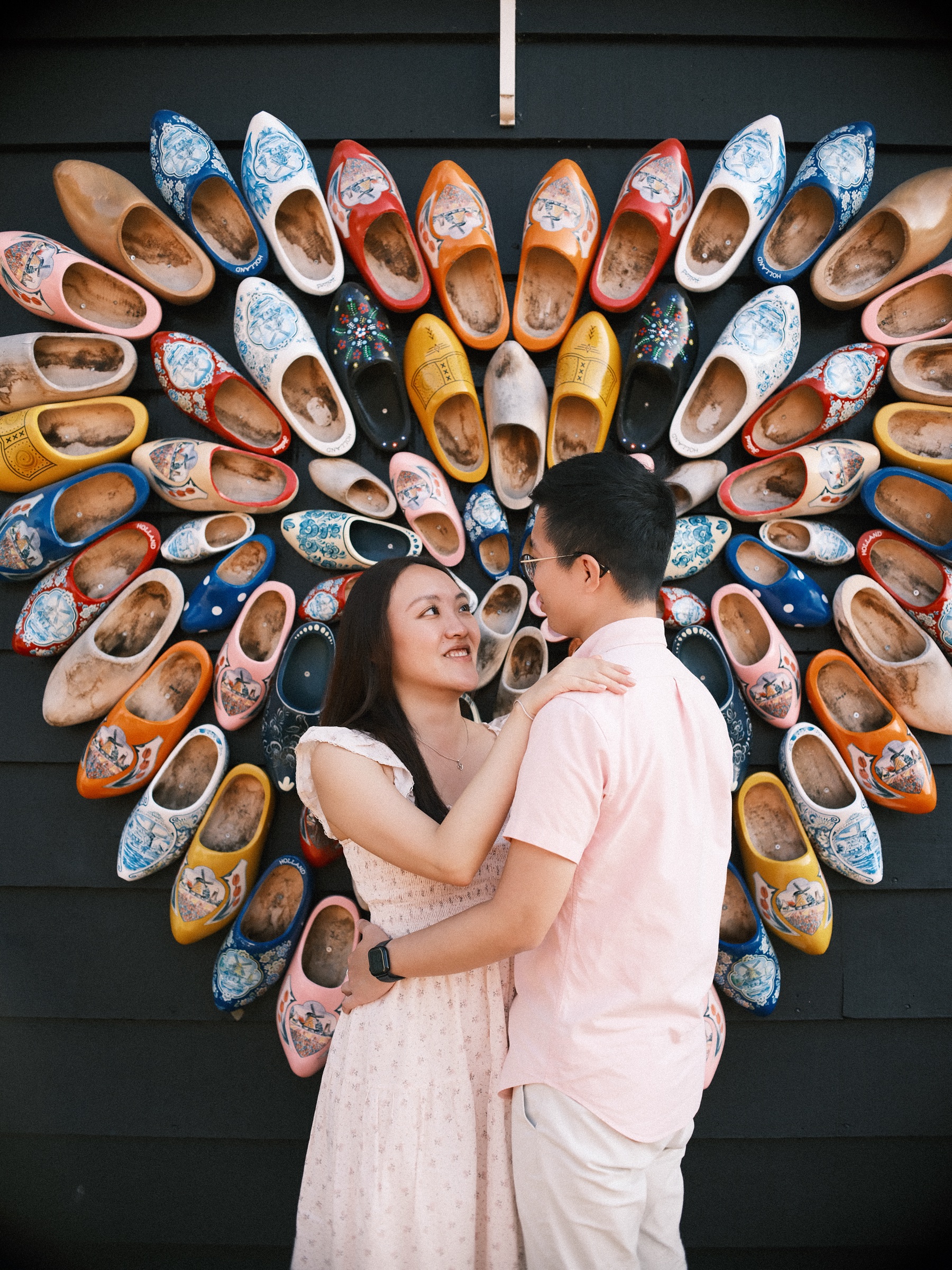 Close embrace before a heart of colorful painted clogs, the woman smiling up at her fiancé, his watch on the hand at her waist