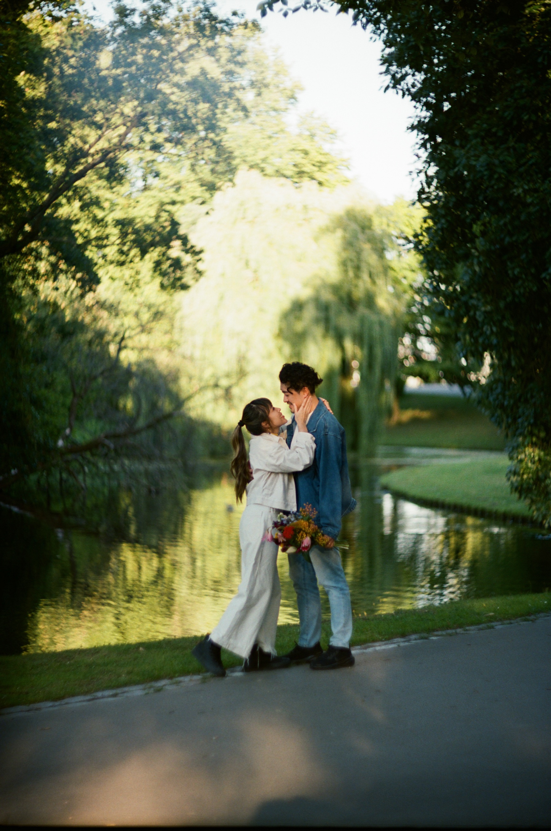 Stel houdt elkaar vast op een pad langs een vijver met treurwilgen, een beeld op Kodak Portra 400 in de avondzon