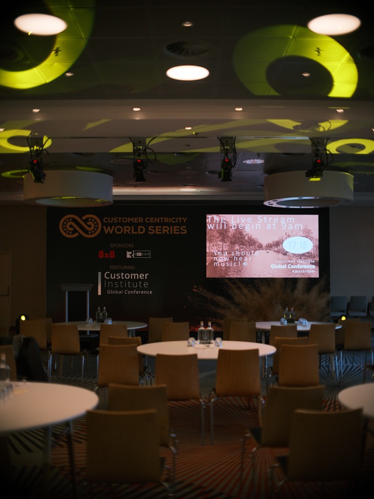 Lege congreszaal met ronde tafels, een sponsorwand en een scherm dat de livestream om negen uur aankondigt
