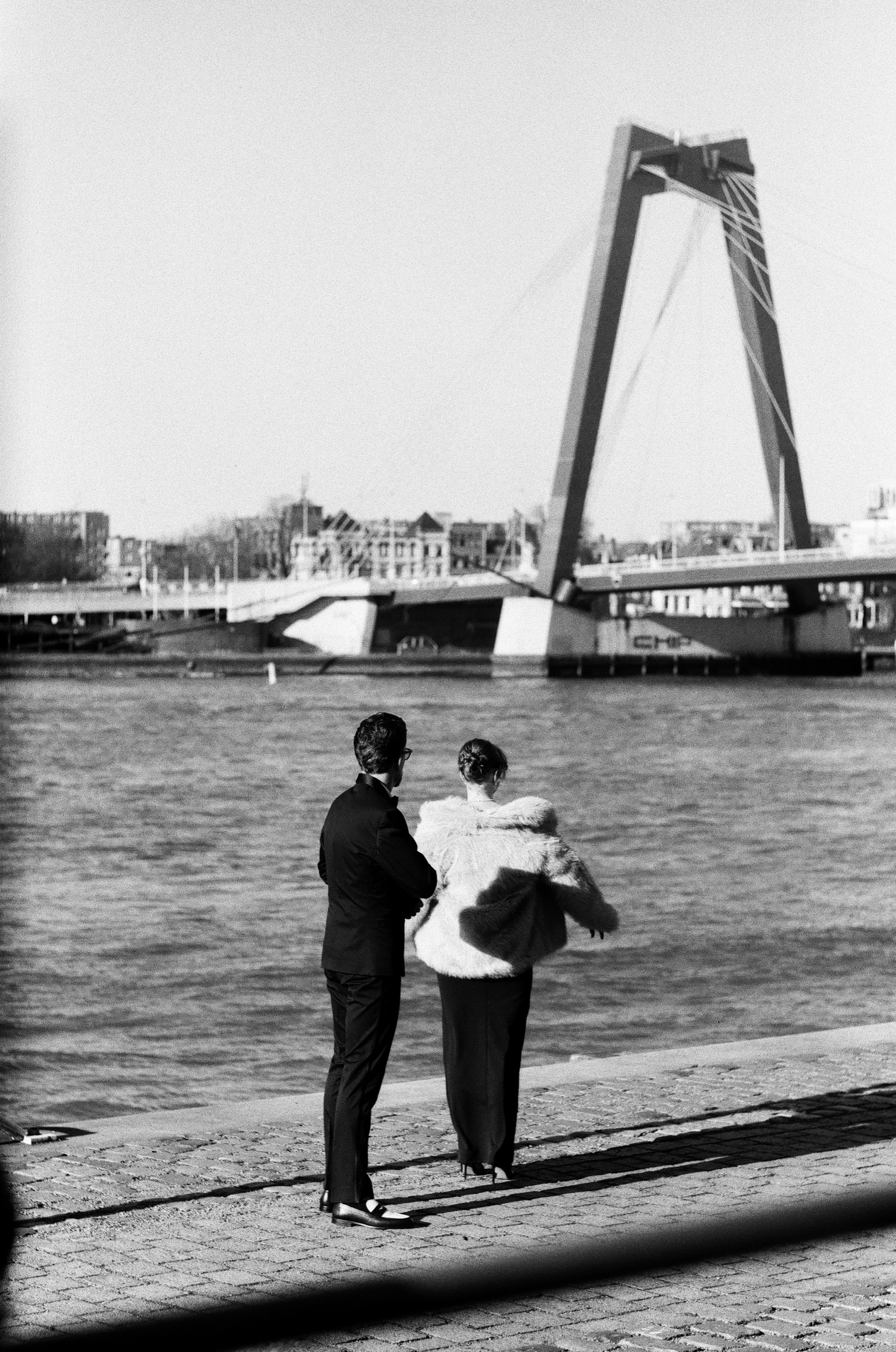 Seen from behind, the newly engaged couple stands arm in arm on the quay, looking at the Willemsbrug after the proposal on film