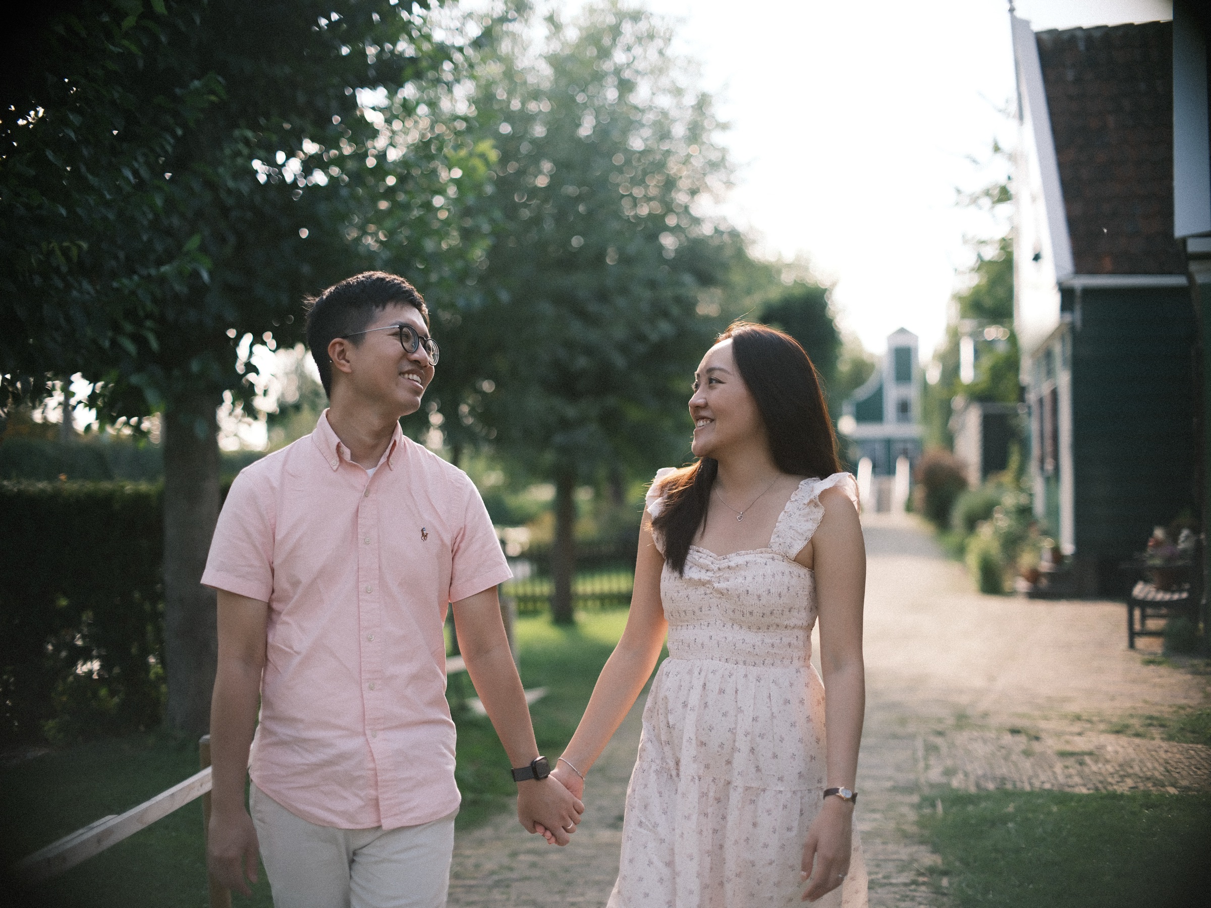 Man in glasses and a pale pink shirt laughing with a woman in a flowered dress as they walk hand in hand through the village