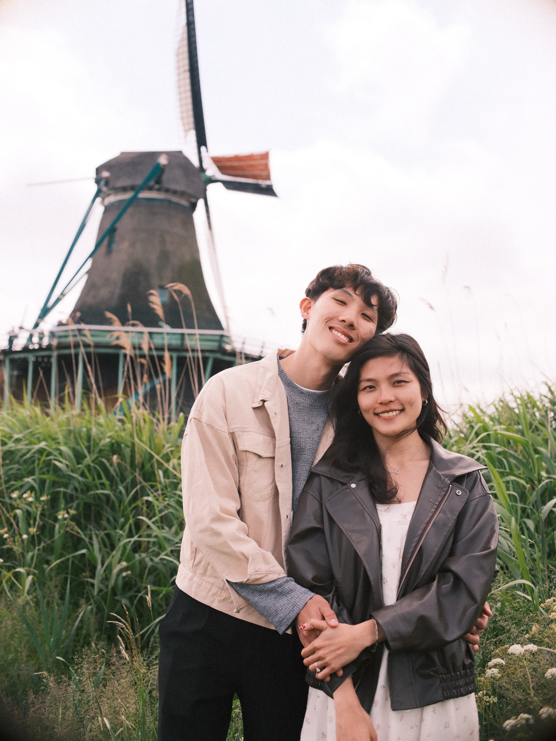 Engaged couple standing close and smiling in tall grass before a dark wooden windmill, just after the proposal at the Zaanse Schans