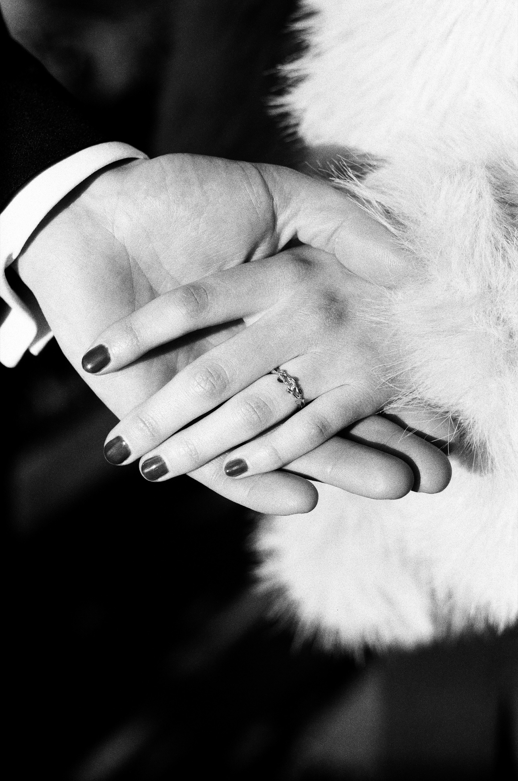 Her hand with a new engagement ring and dark nails resting on his hand, the soft fur of her coat behind, shot on black and white film