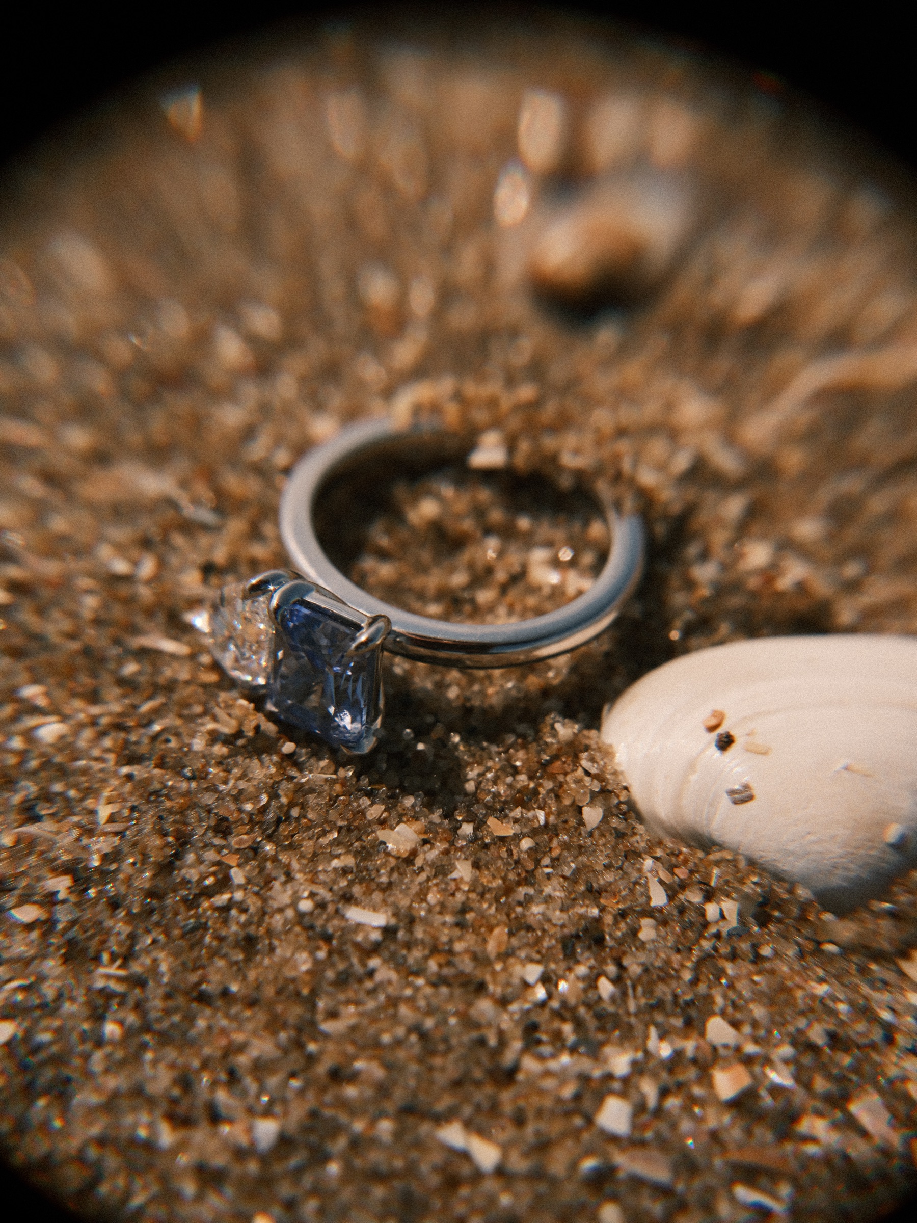 Engagement ring with a blue and a clear stone lying on the sand beside a white shell, soft swirling blur at the edges