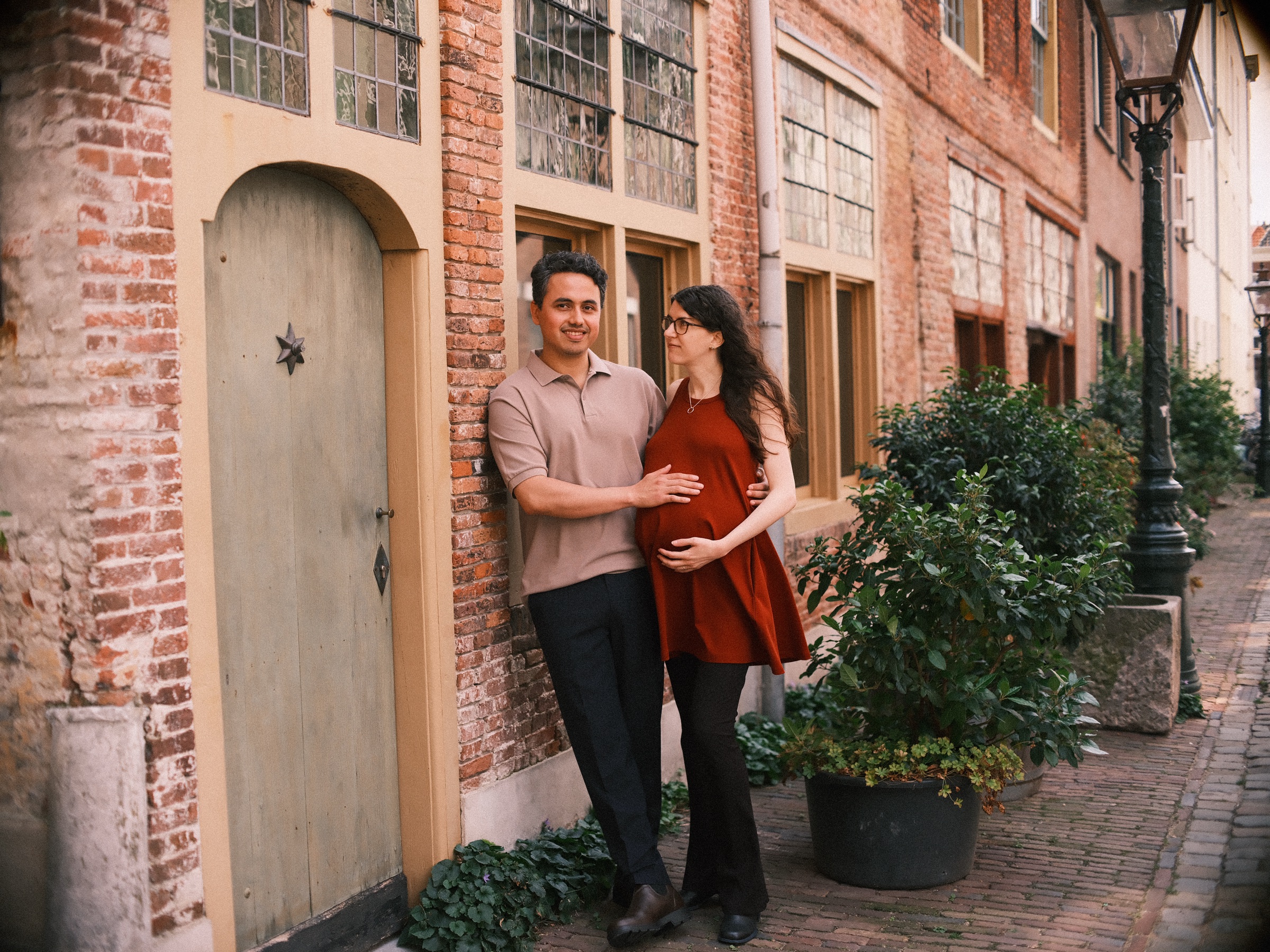 Expecting couple by a pale arched door with a small star, he in a taupe polo after a change of outfit, plants and an old lamppost beside them